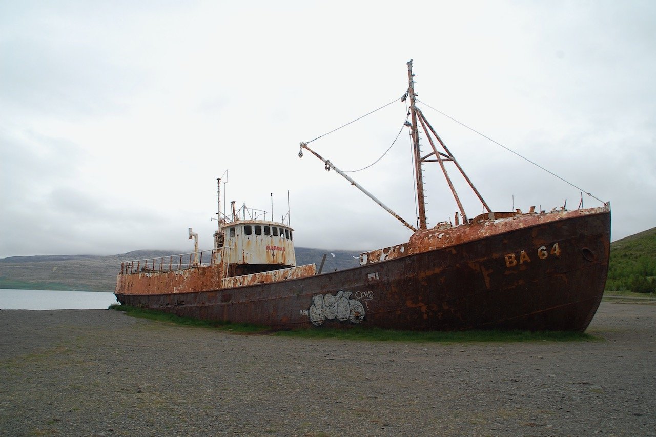 A photo of a large rusted ship that has come aground on a gravelly shore. The sky is a bleached out gray. Photo credit: Kelly Nielsen