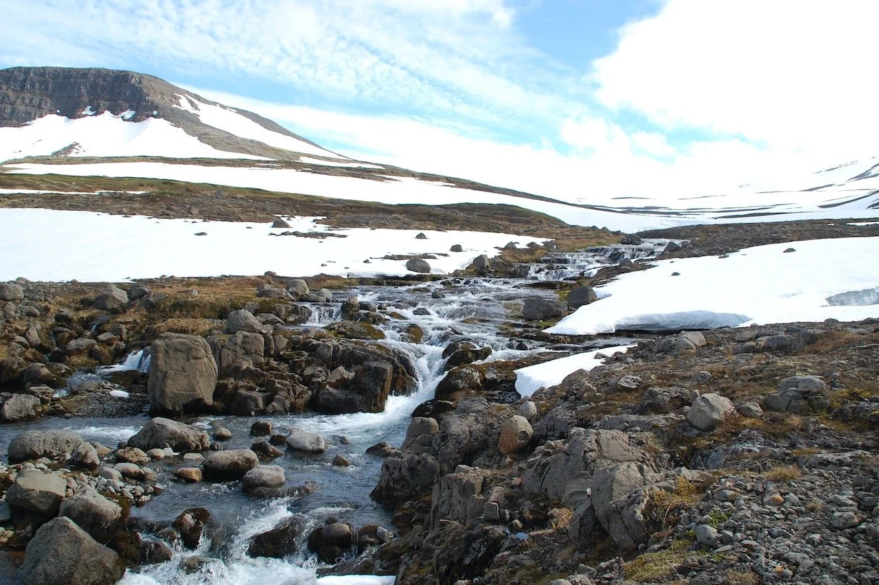A landscape photo of large rocks and snow with a rocky hill visible in the distant left. A rocky stream flows toward the camera. Taken in Iceland. Photo credit: Kelly Nielsen