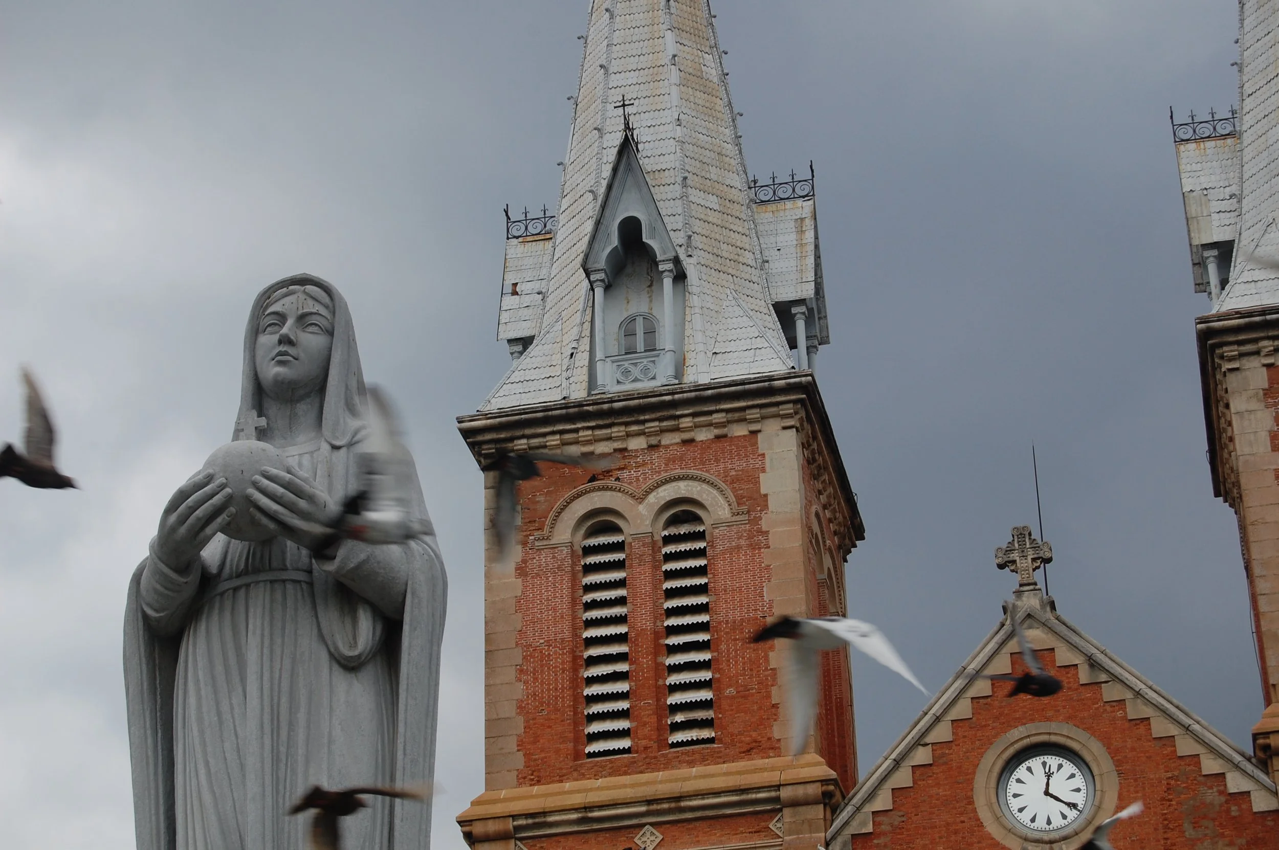 In the left of the photo is a statue of a veiled woman, likely Mary. In the center and right of the photo are the steeple and clock of a red brick church. Birds fly across the frame in a blur with a dark sky behind. Photo credit: Kelly Nielsen