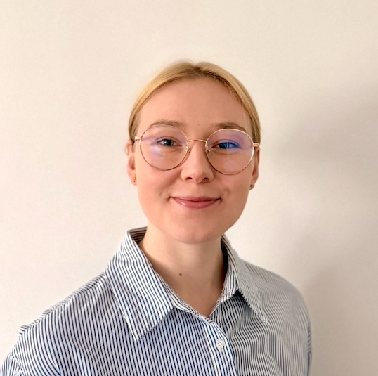 Une jeune femme avec des lunettes rondes, des cheveux blonds attachés, portant une chemise à rayures, souriant devant un mur blanc.