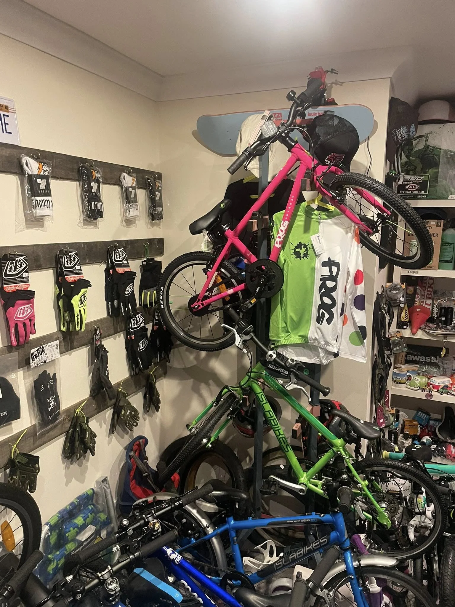 A room with various bicycles, a pink bike mounted on a wall rack, mountain bikes, bicycle accessories, and shelves with toys and collectibles at the Thee54 Bike Store