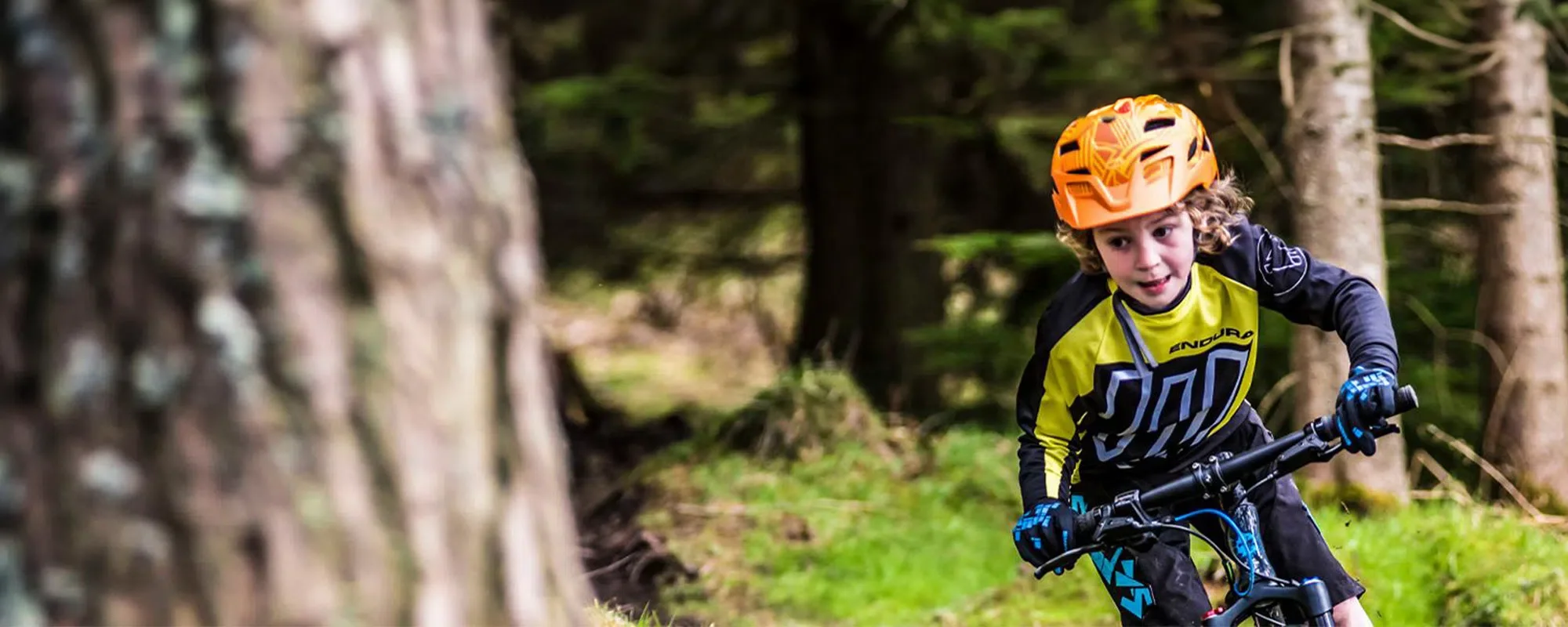 Child mountain biking through a forest trail, wearing an orange helmet and yellow sports jersey. Hurrying to get to Three54 Bike Store