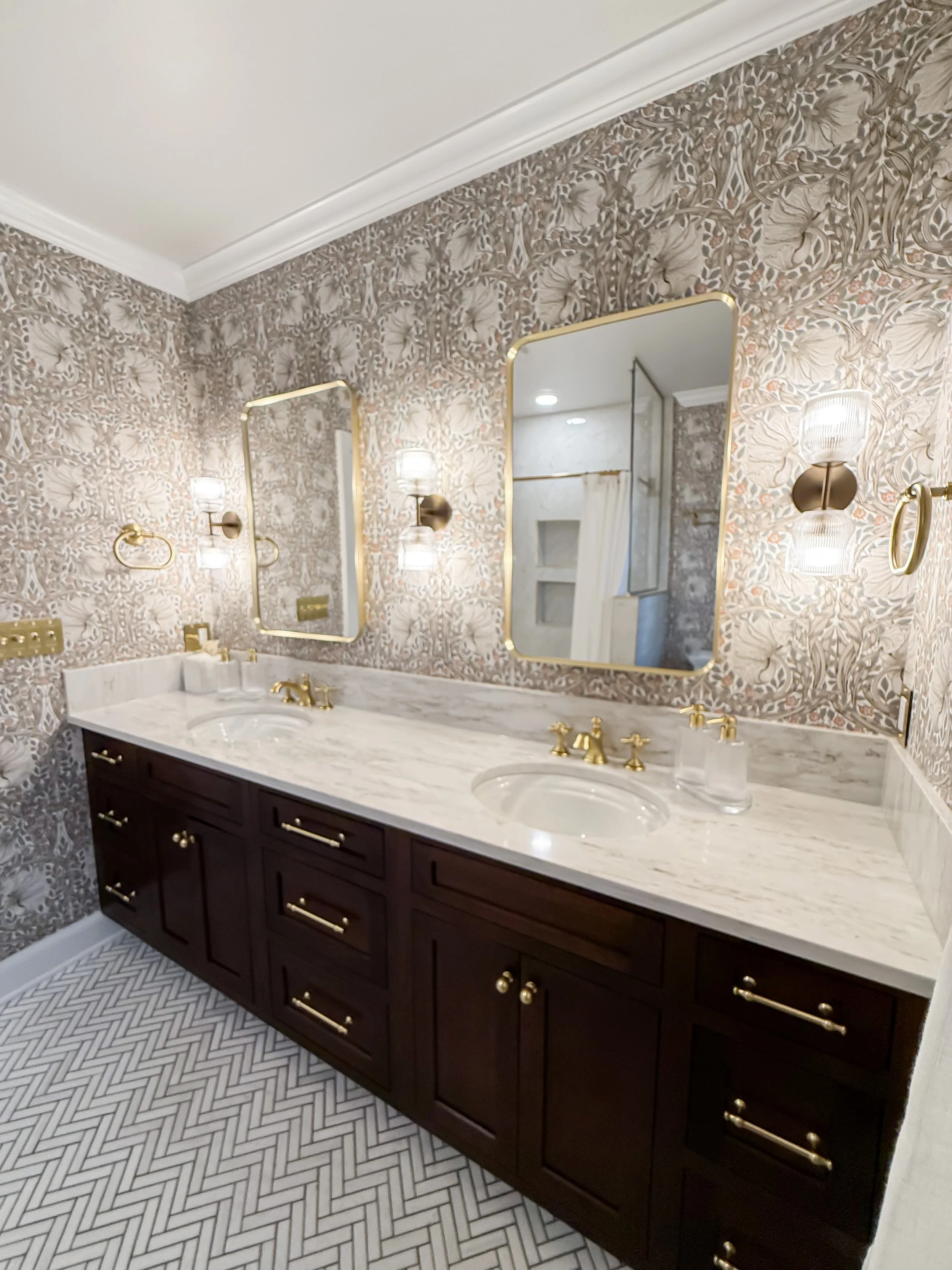 A double-sink bathroom vanity with a marble countertop, dark wood cabinet, gold hardware, two mirrors, and decorative lighting fixtures on a floral wallpaper wall.