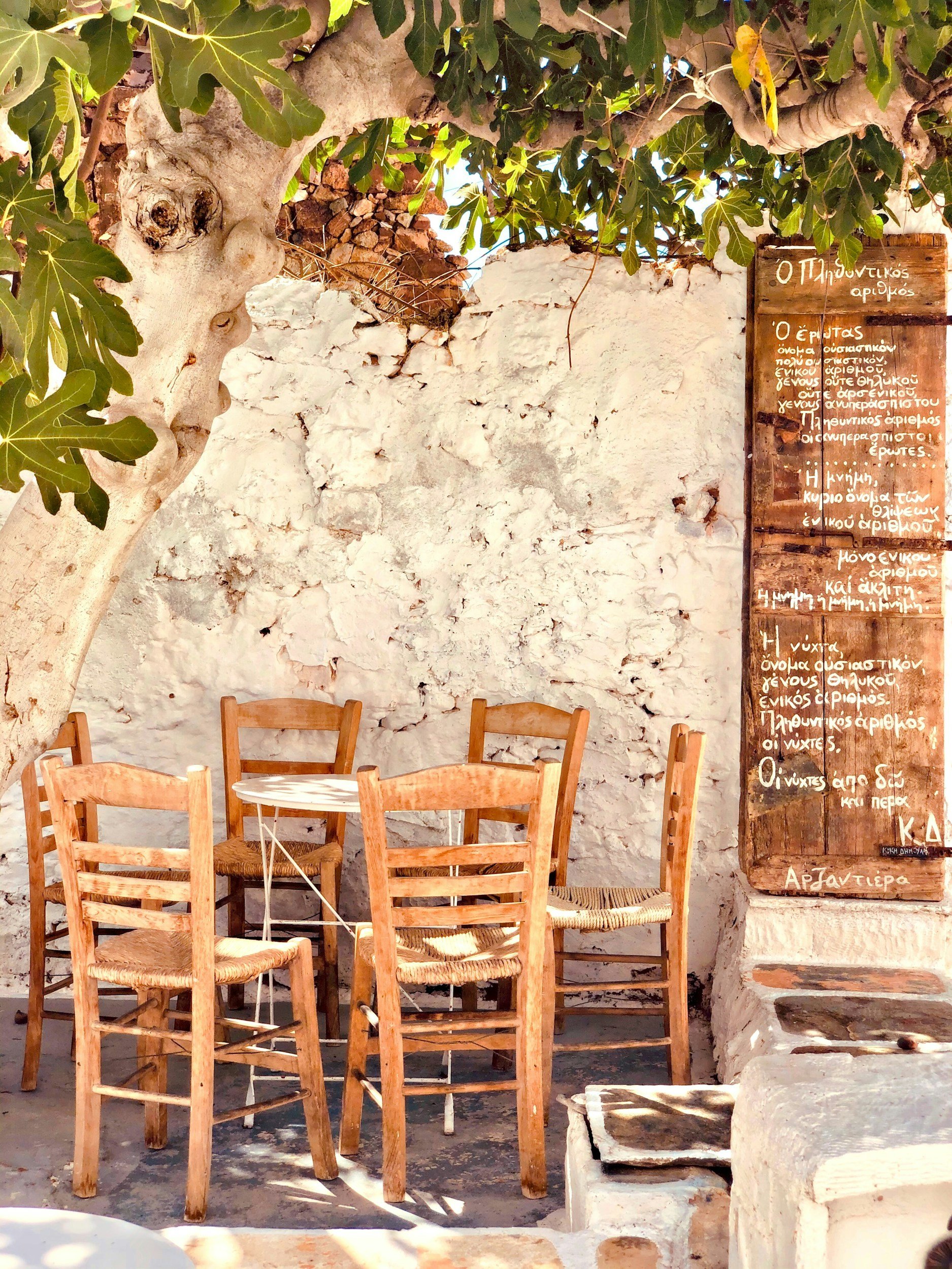 A cozy outdoor seating area with five wooden chairs around a small round table, set against a white textured stone wall with a wooden signboard with Greek text on the right side and a leafy tree providing shade.