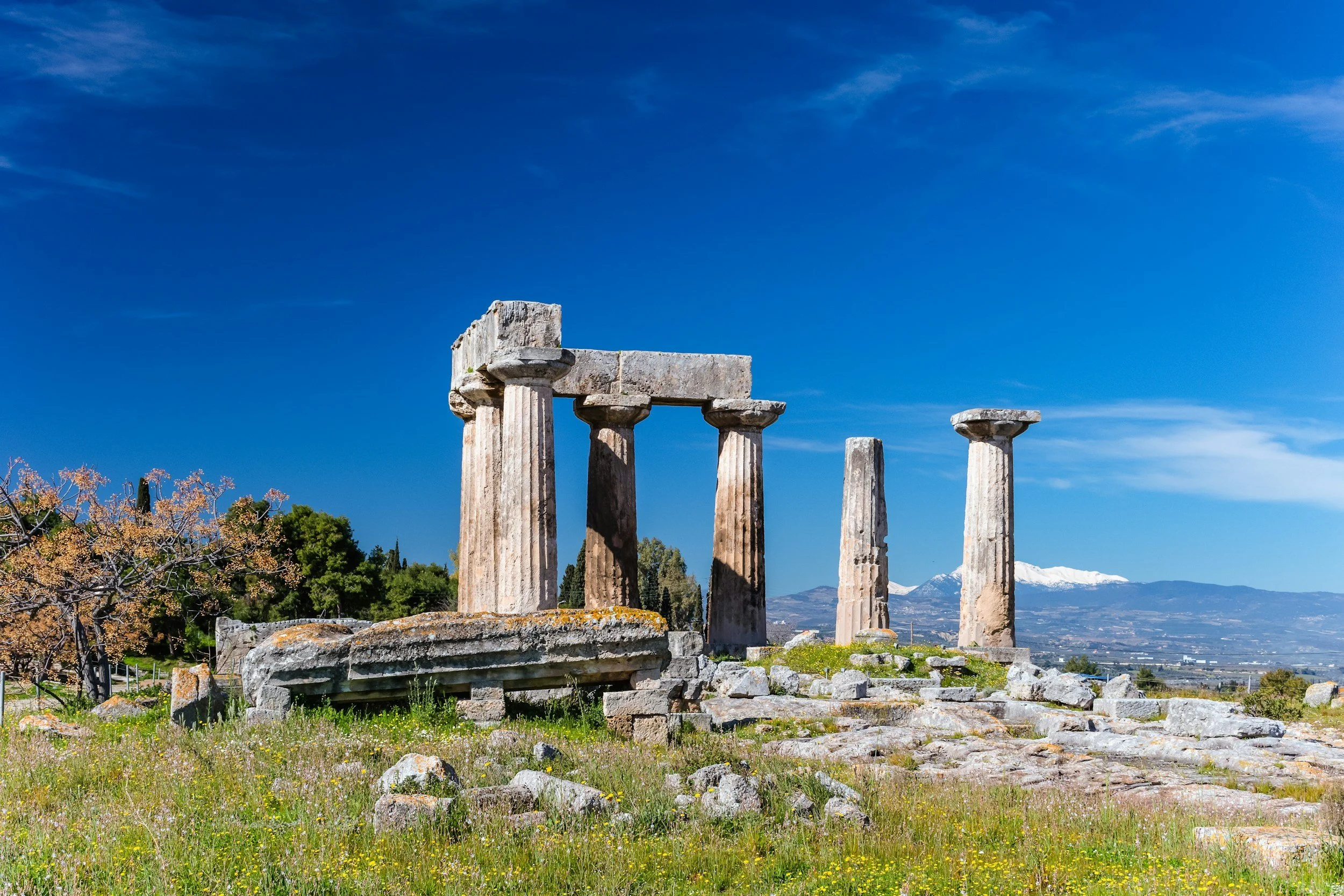 Ancient Greek ruins with four standing columns and one lying horizontal, set against a bright blue sky with some clouds and snow-capped mountains in the background.
