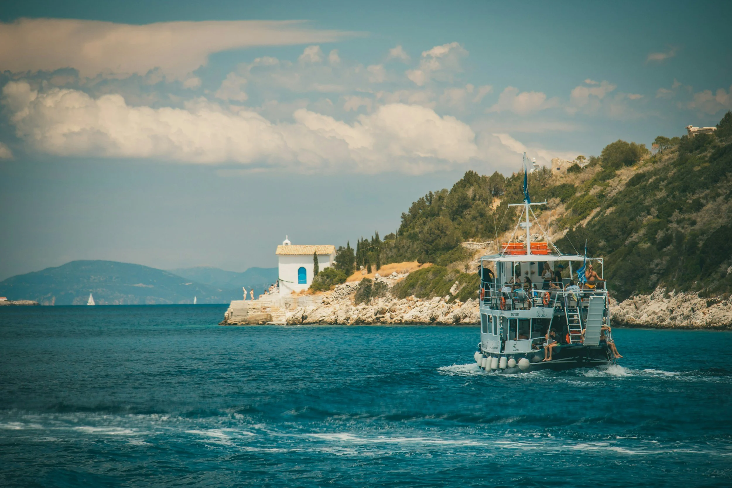 A boat on the water near a rocky coastline with a small white building with a blue window and door, hills, and a partly cloudy sky in the background.