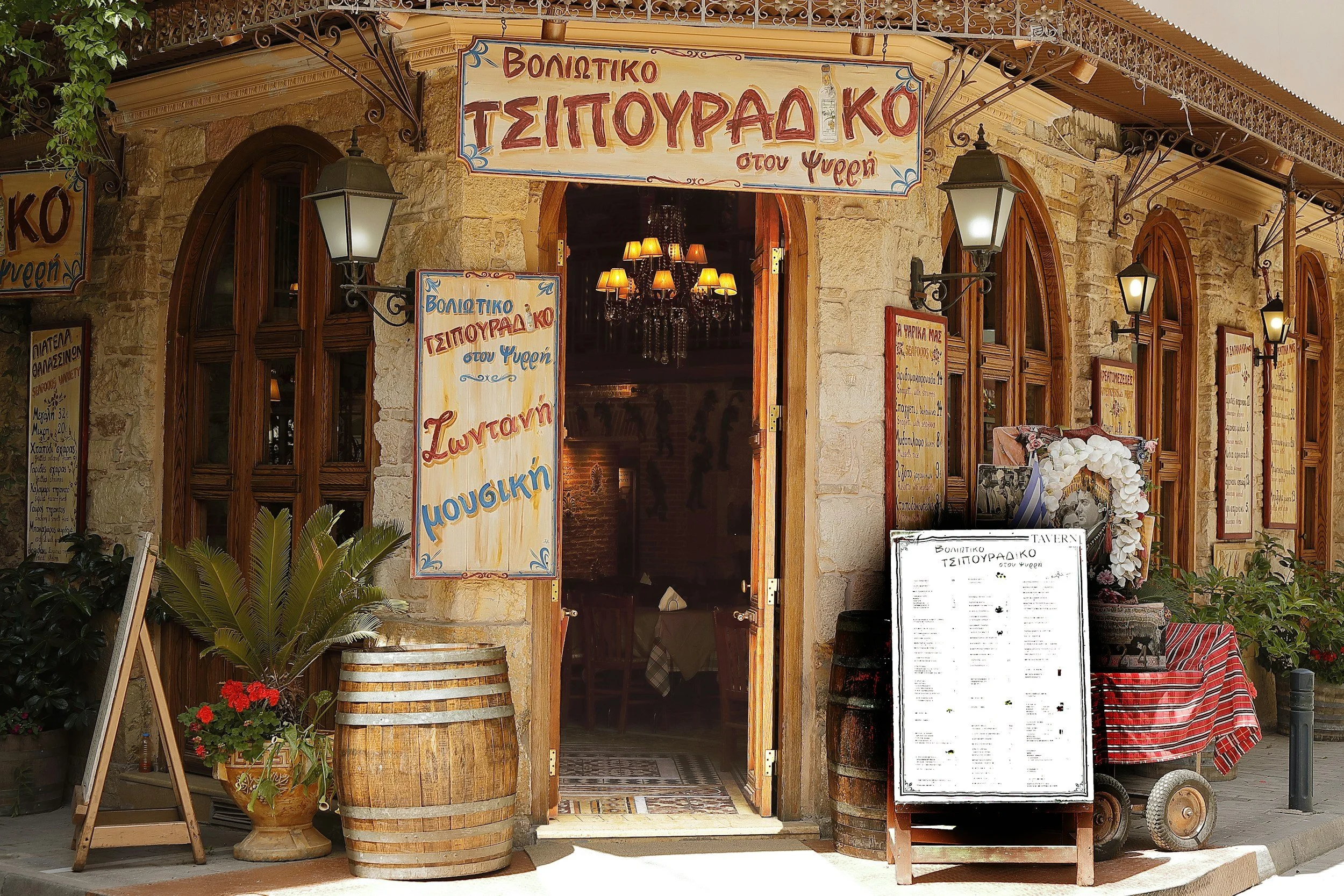 Entrance of a Greek restaurant with wooden doors, stone walls, and Greek signs. There are potted plants, a barrel, a chalkboard menu, and decorative lighting fixtures.