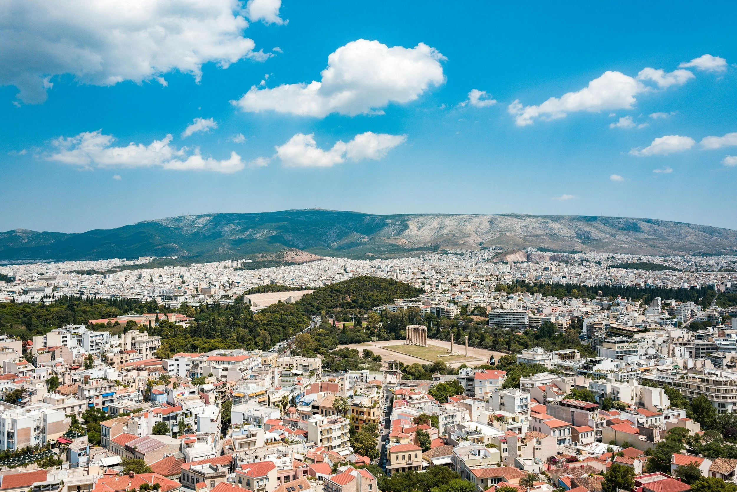 A panoramic view of a city with white buildings, green parks, and a distant mountain range under a partly cloudy blue sky.