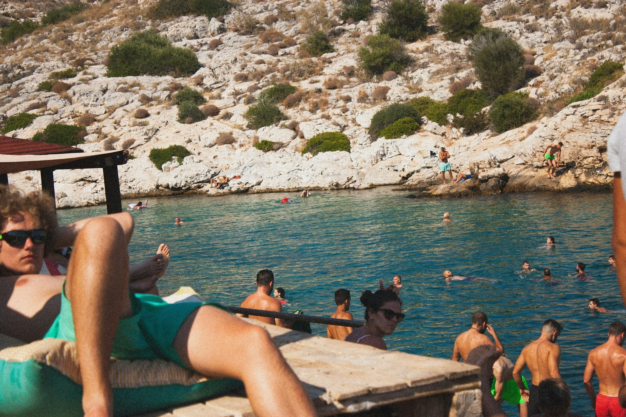 People swimming and relaxing near a rocky shoreline by a body of water with a hill covered in rocks and sparse vegetation in the background.