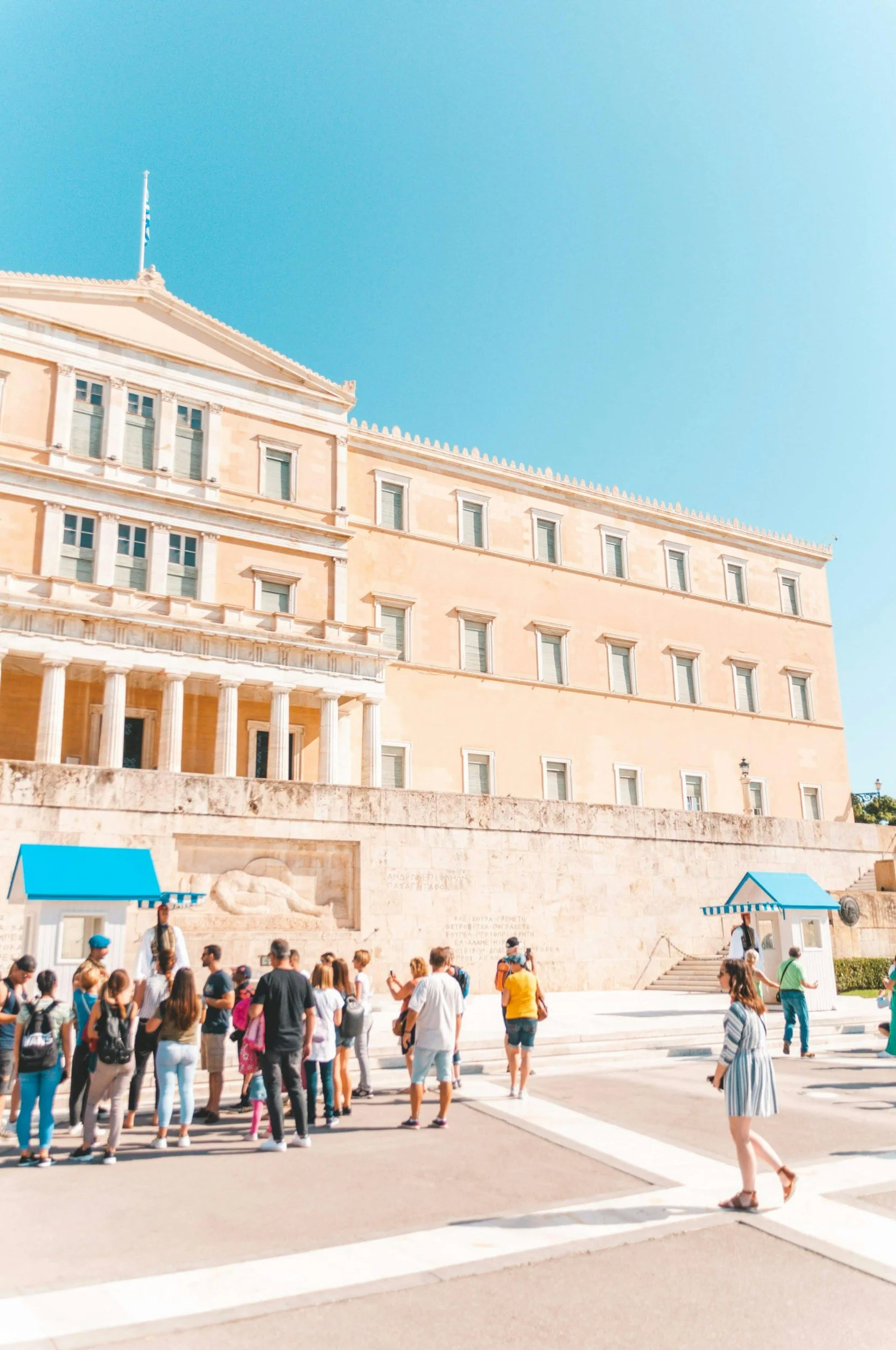 A group of tourists standing outside the Greek Parliament building in Athens, Greece, with the Hellenic Parliament behind them, featuring classical architecture and a flag on top, under a clear blue sky.
