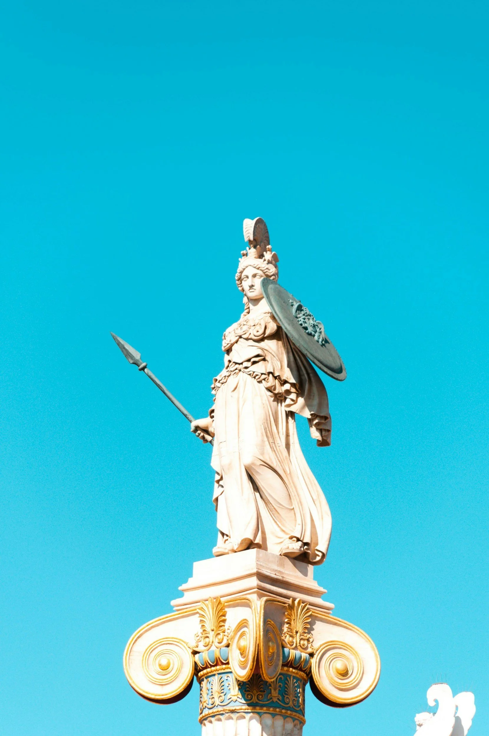 A classical statue of a woman dressed in ancient armor, holding a spear and shield, standing on an ornately decorated column with a clear blue sky in the background.