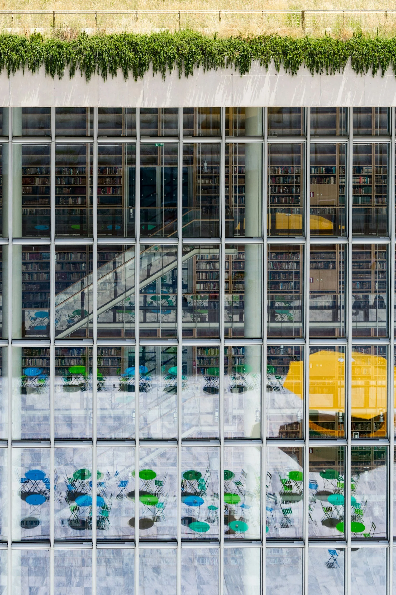 A glass building with a library inside, showing bookshelves, colorful chairs, and tables viewed through the windows. The upper section has a rooftop garden with green plants.