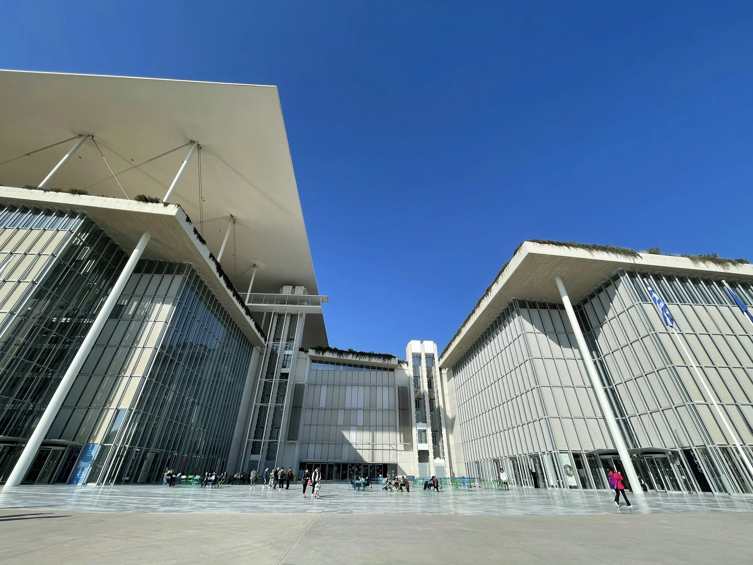 Modern building with large glass windows and white architectural features, facing a spacious outdoor plaza with people walking, under a clear blue sky.