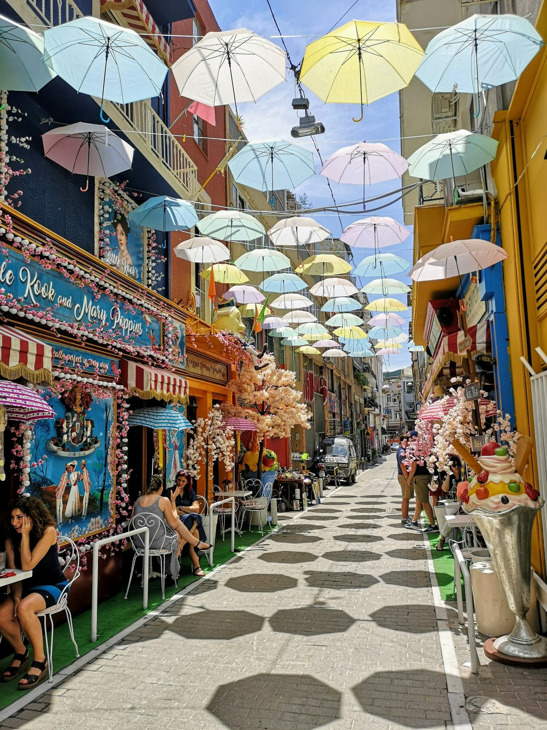 Colorful street decorated with hanging umbrellas and pink cherry blossom decor, with people sitting and walking.