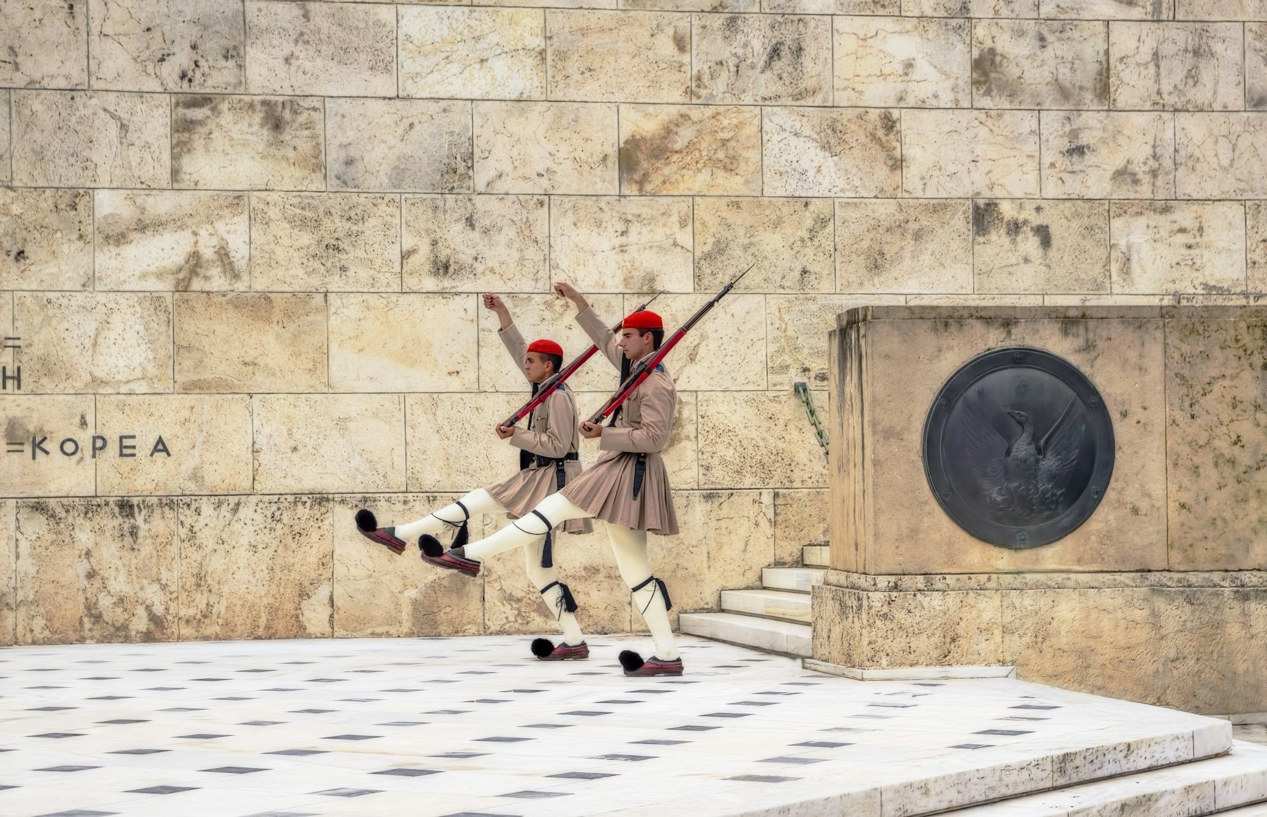 Two traditional guards in tan uniforms and red hats marching with rifles, raising one arm in a salute, on a black and white tiled ground against a stone wall with a commemorative plaque.