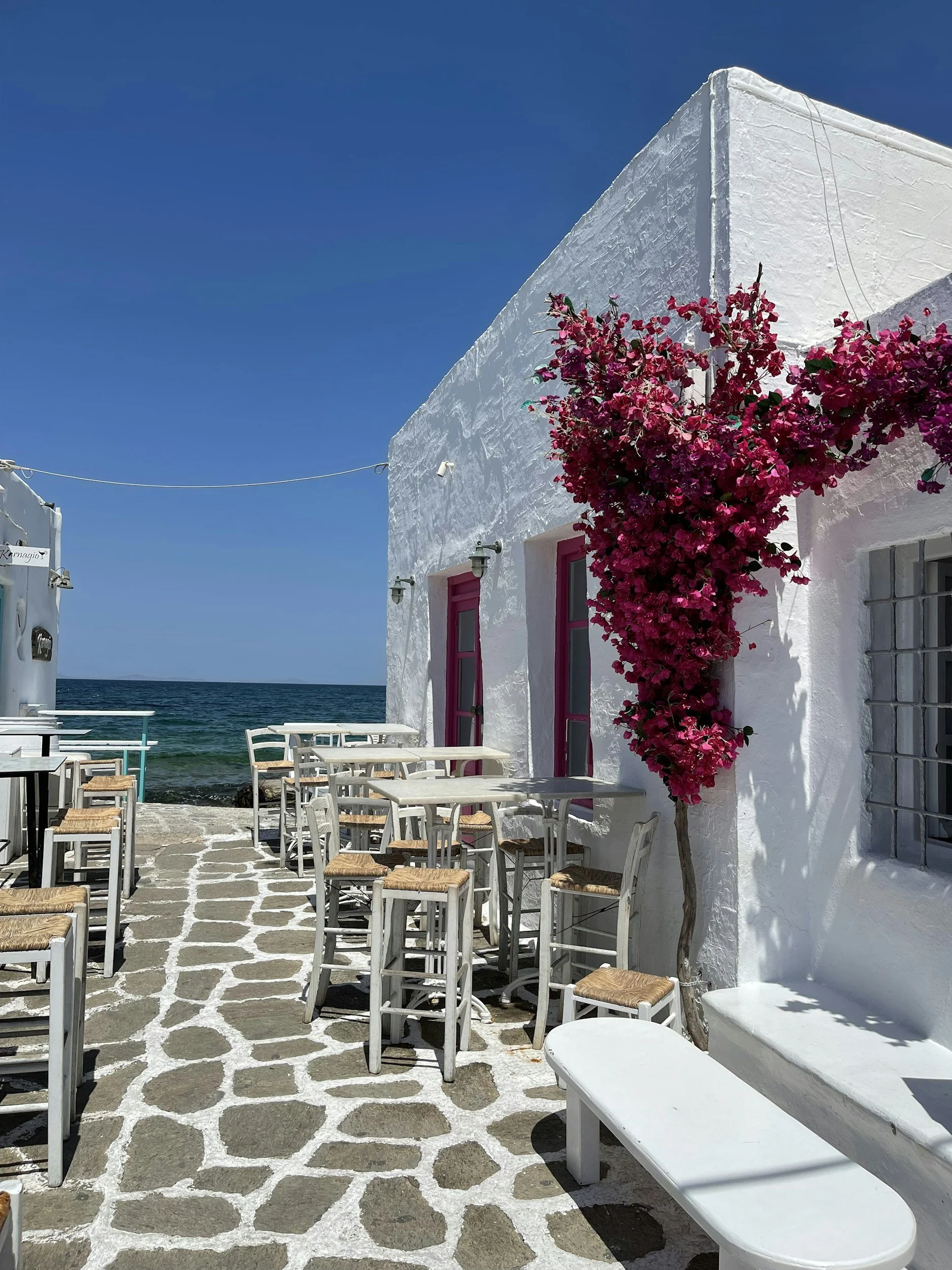 White building with pink window frames and bougainvillea climbing the wall, overlooking the sea, with outdoor seating and a clear blue sky.