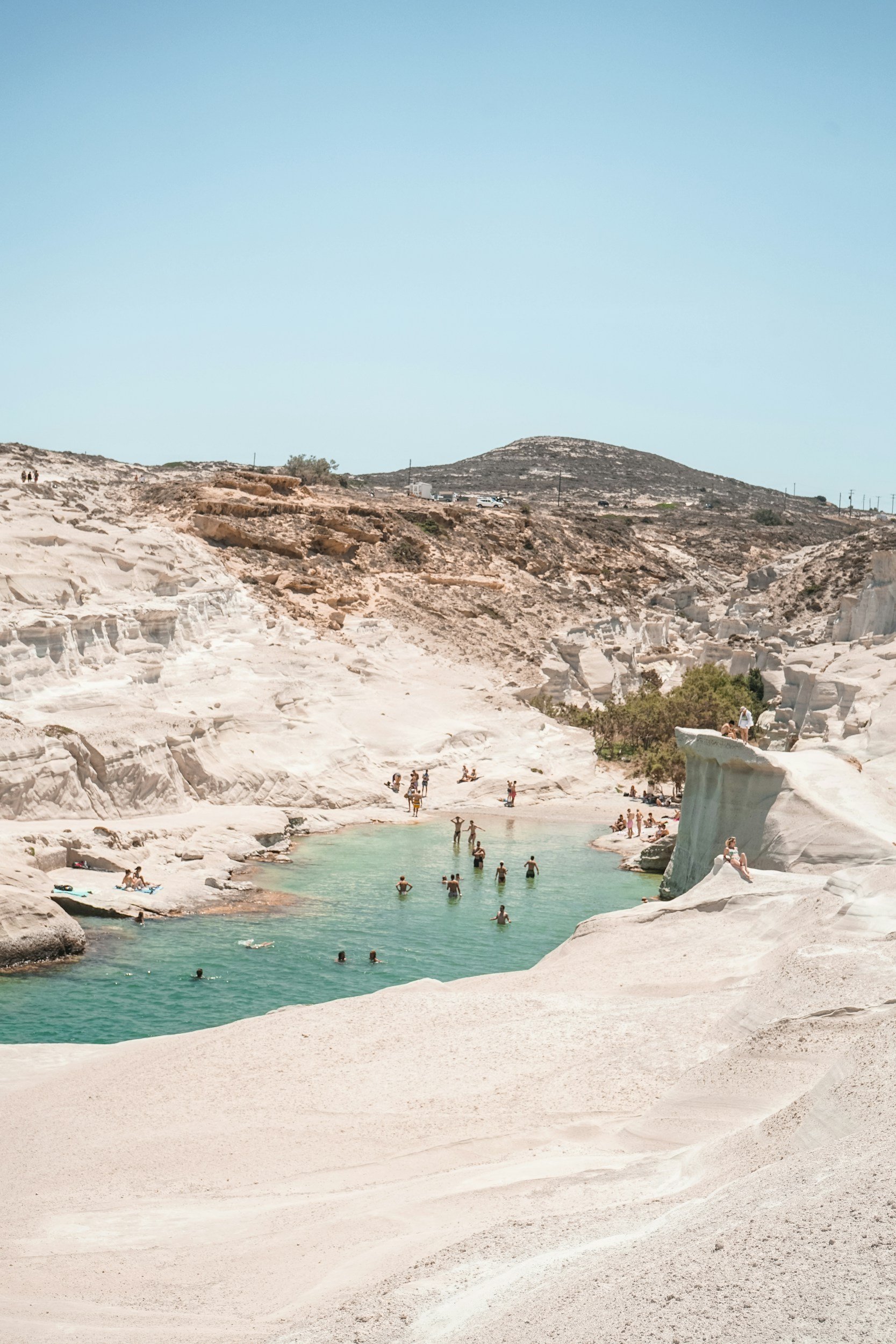 People swimming and relaxing in a natural hot spring surrounded by white rock formations, with dry, rocky hills in the background under a clear sky.