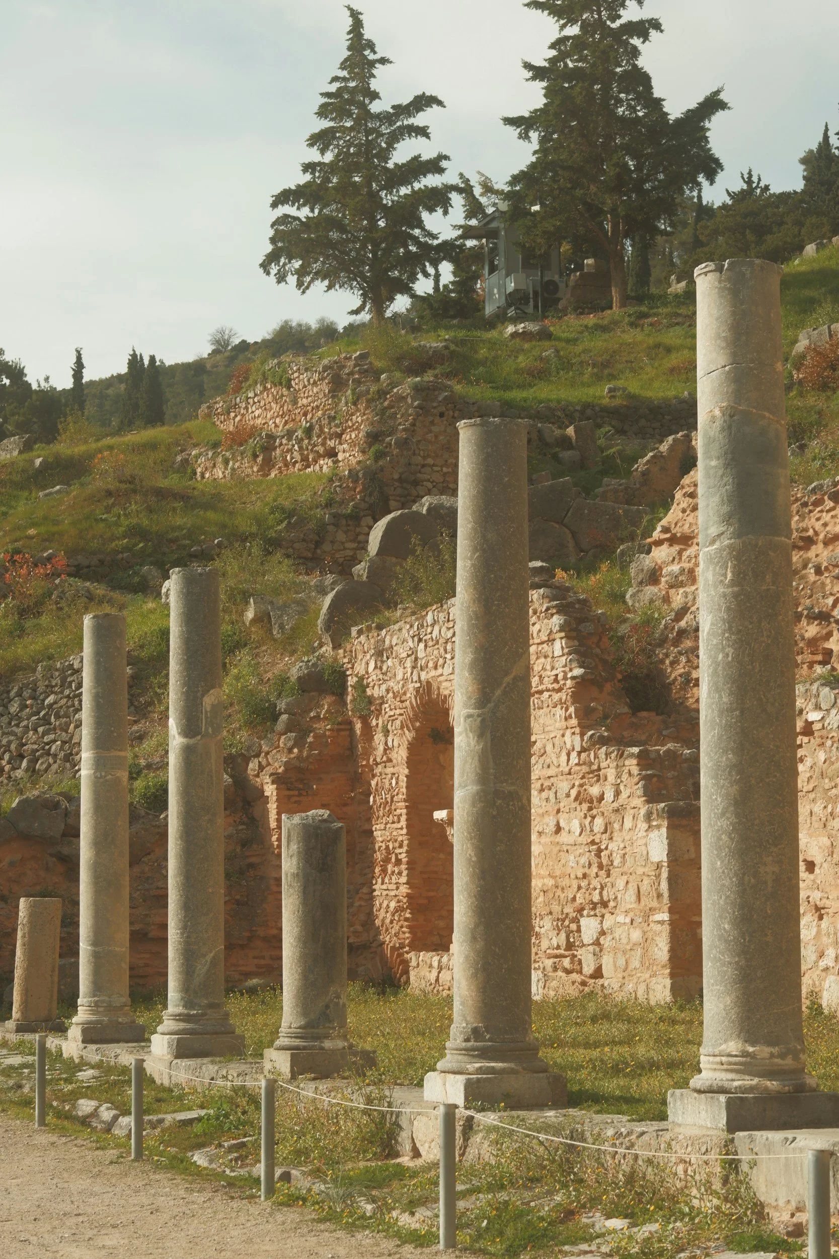 Ancient stone ruins with stone columns and brick walls, situated on a grassy hillside with trees, under a cloudy sky.