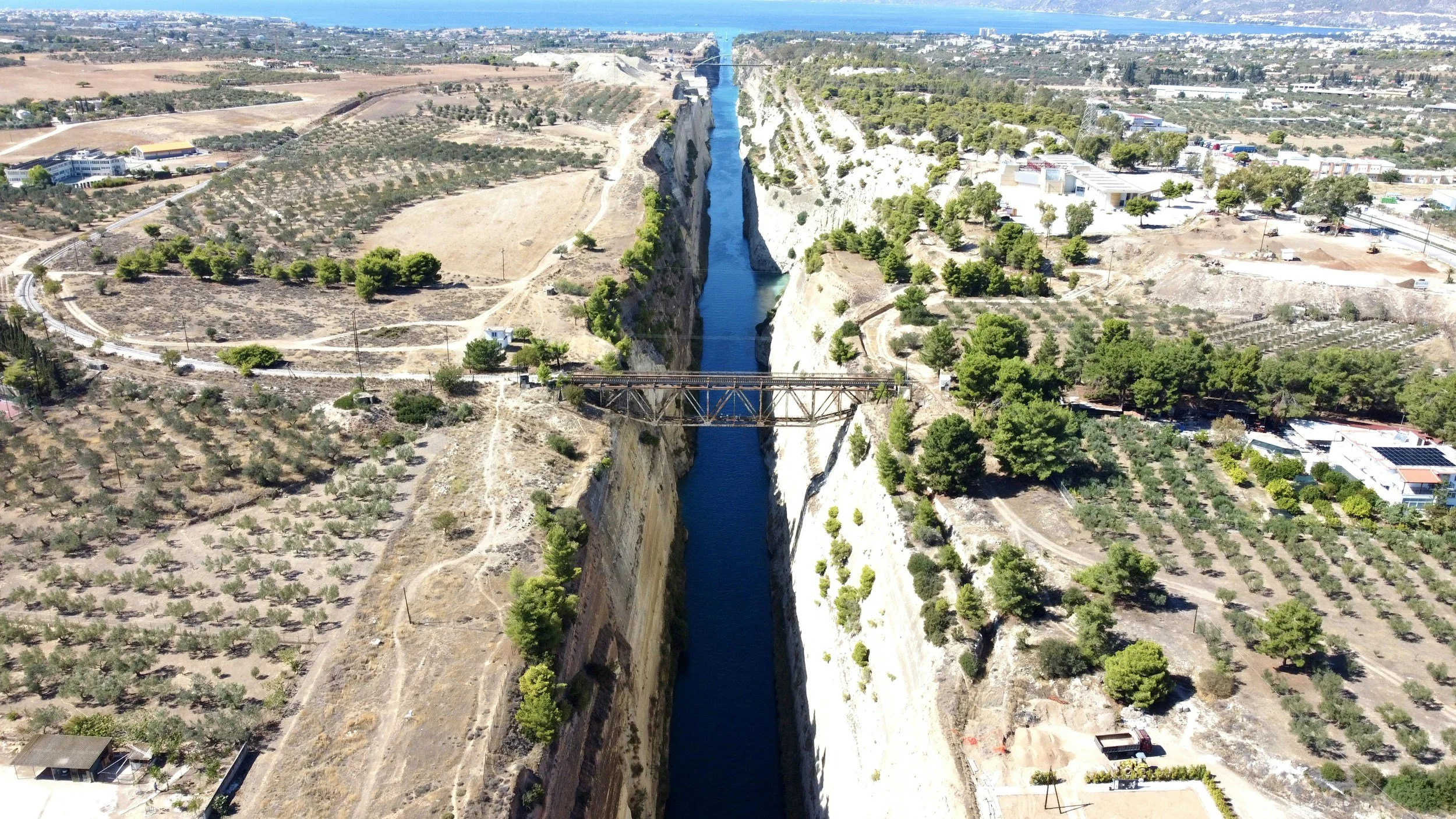 An aerial view of a large canyon with steep white cliffs and a river flowing through the bottom. A bridge crosses the canyon, connecting the two sides. There are trees and dry land surrounding the canyon, with some buildings and roads visible in the distance.