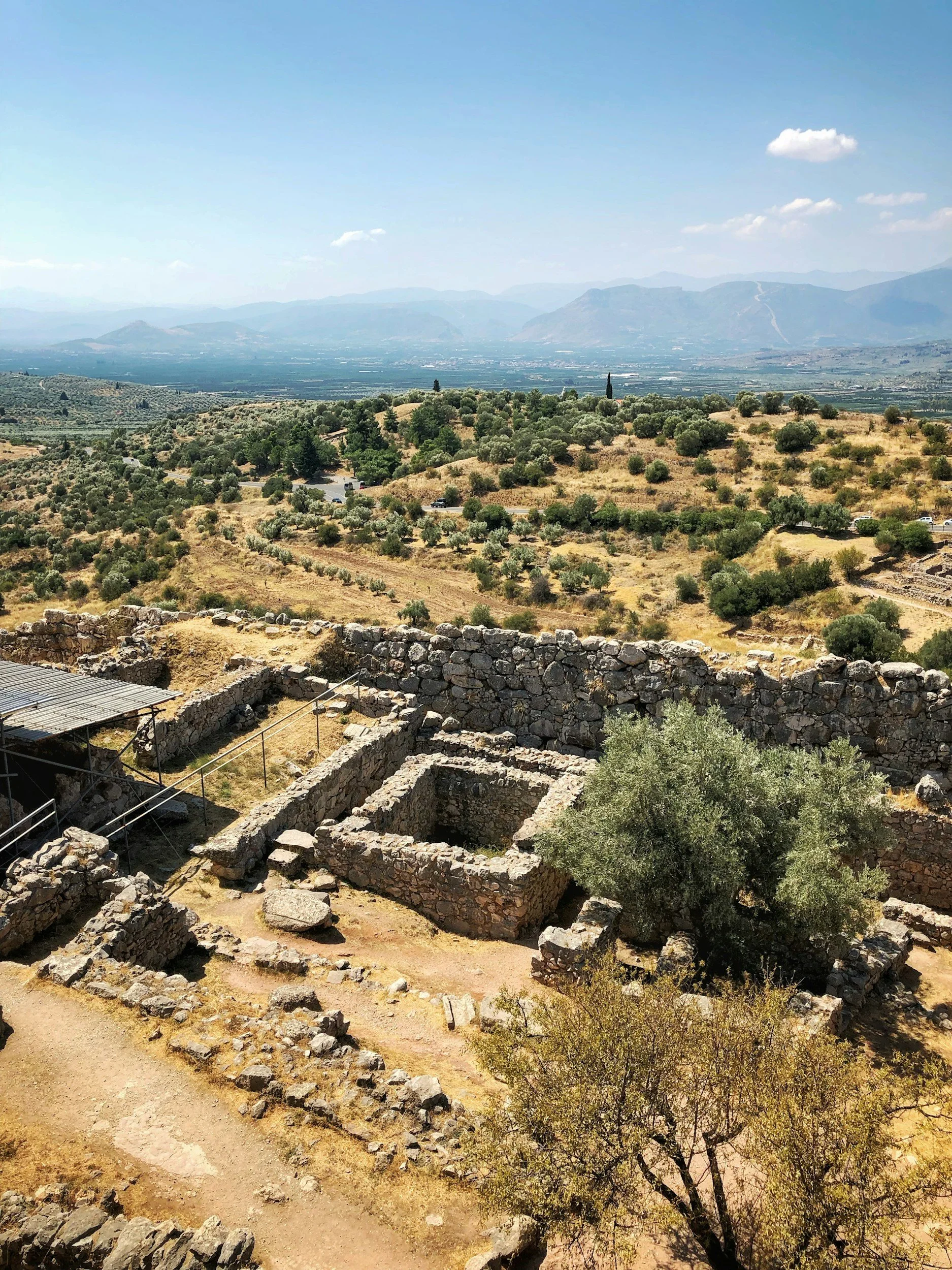 Ruins of an archaeological site with stone walls and a dirt pathway, set in a hilly landscape with scattered trees and distant mountains under a blue sky with a few clouds.