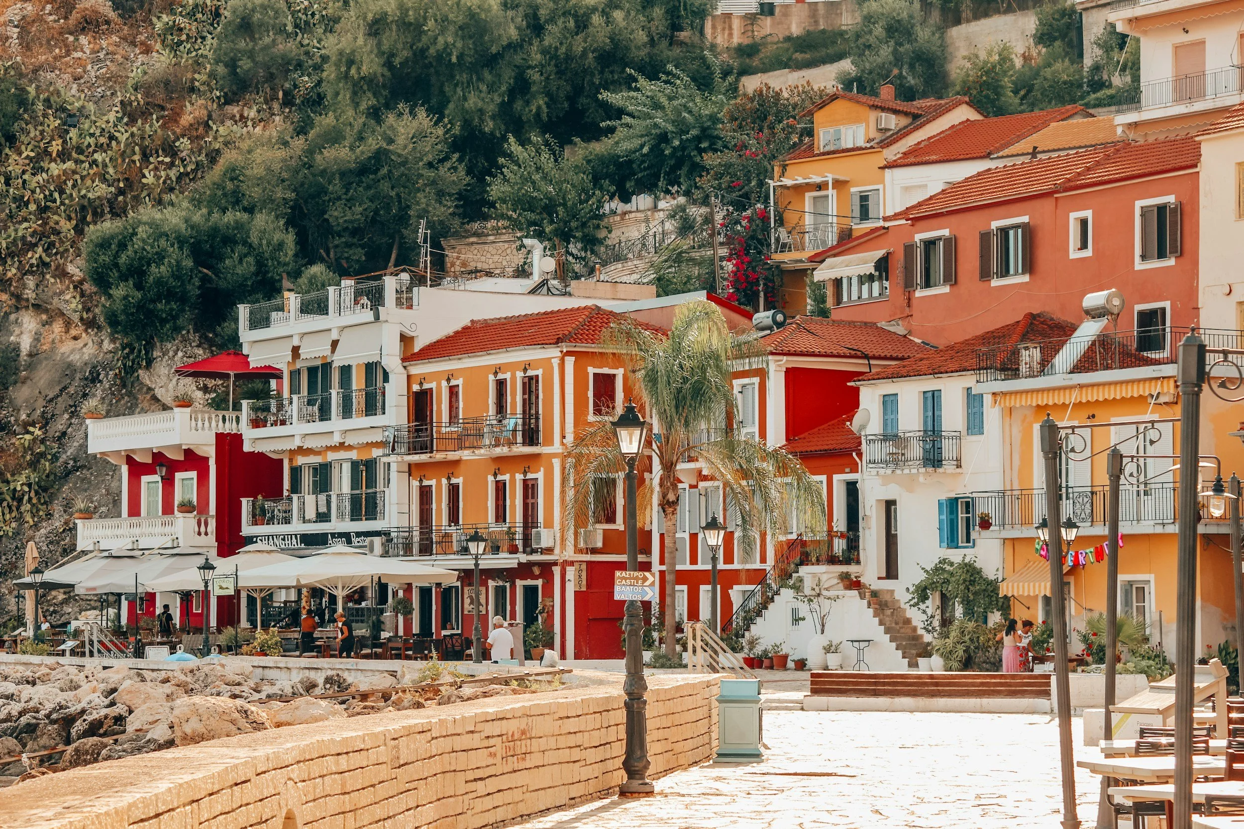 Colorful buildings with orange and white facades, red-tiled roofs, and balconies, set against a hillside with lush greenery, on a sunny day in a coastal town.