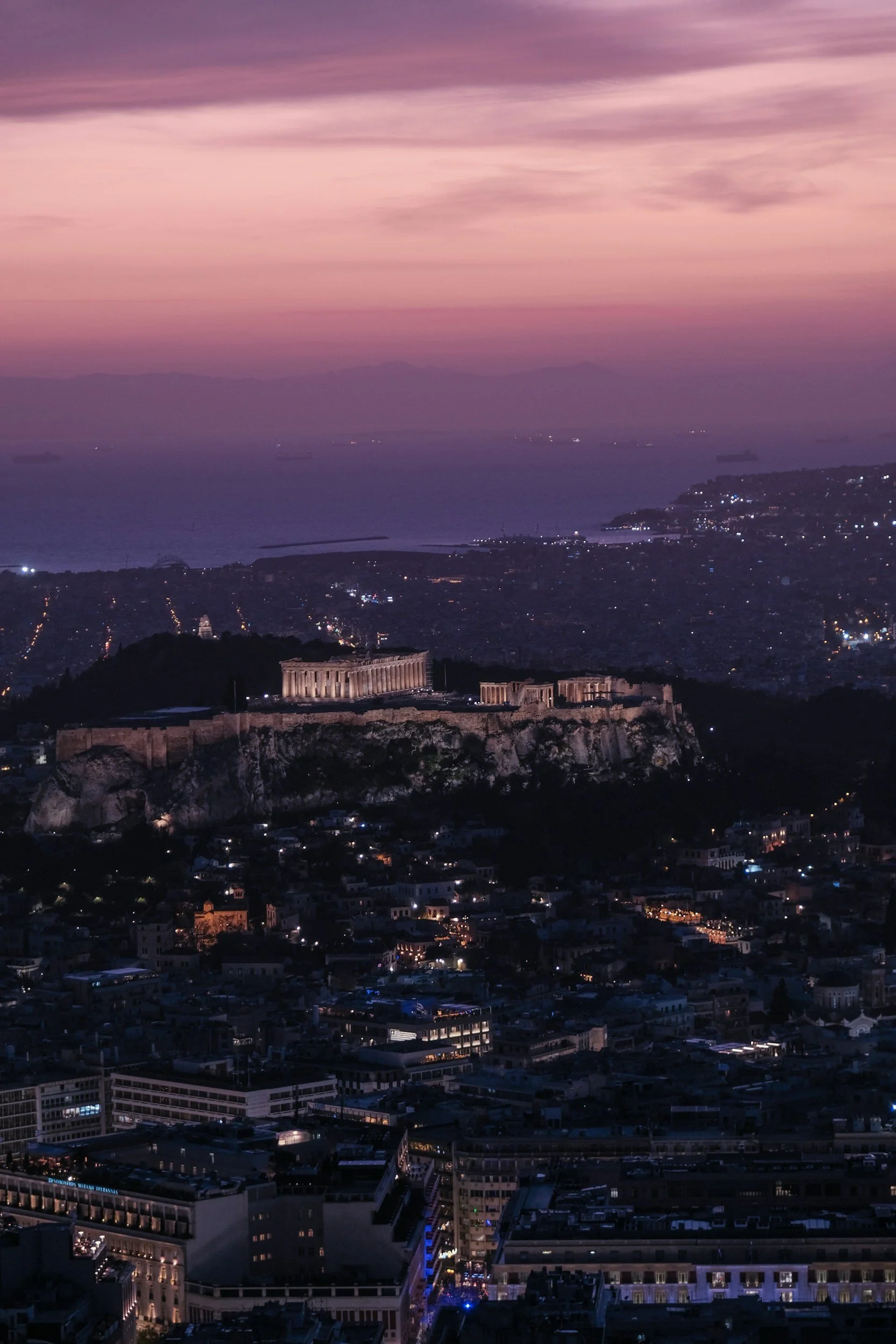 A cityscape at dusk with the Acropolis and Parthenon illuminated on a hill, overlooking a city with numerous buildings and lights, under a purple and pink sky.
