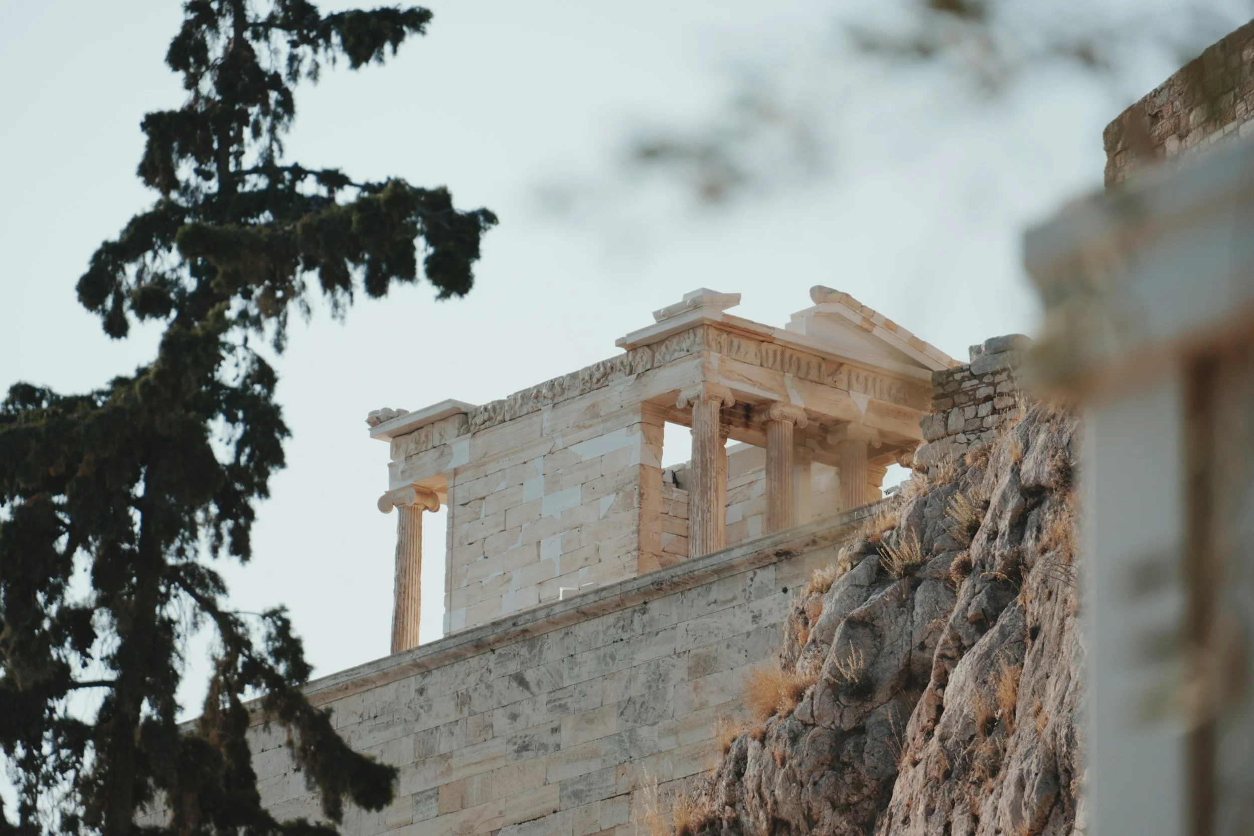 Ancient Greek temple ruins on a rocky hillside with a tree in the background.