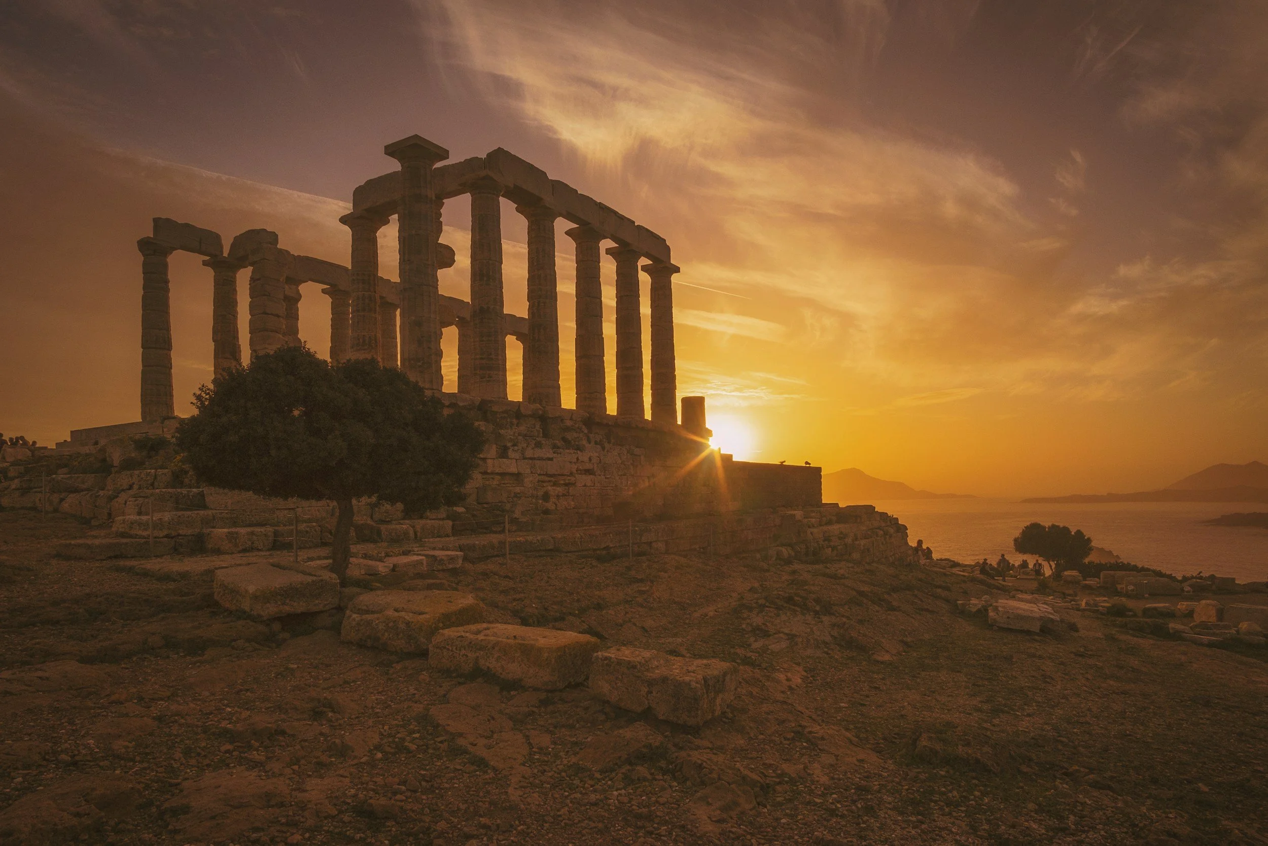 Ancient Greek ruins with tall columns at sunset, overlooking the sea with a vibrant orange sky.