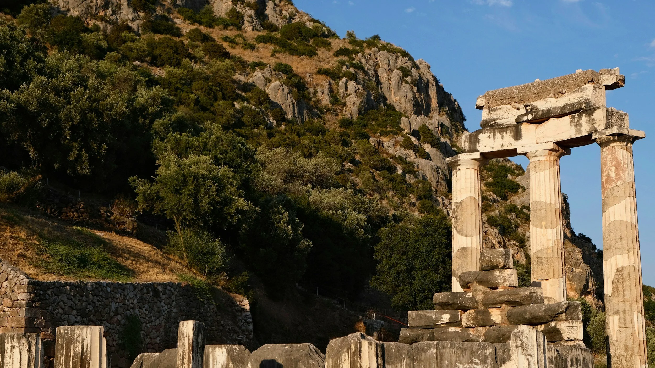 Ancient ruins with three tall stone columns against a hillside covered in trees and rocks, under a clear blue sky.