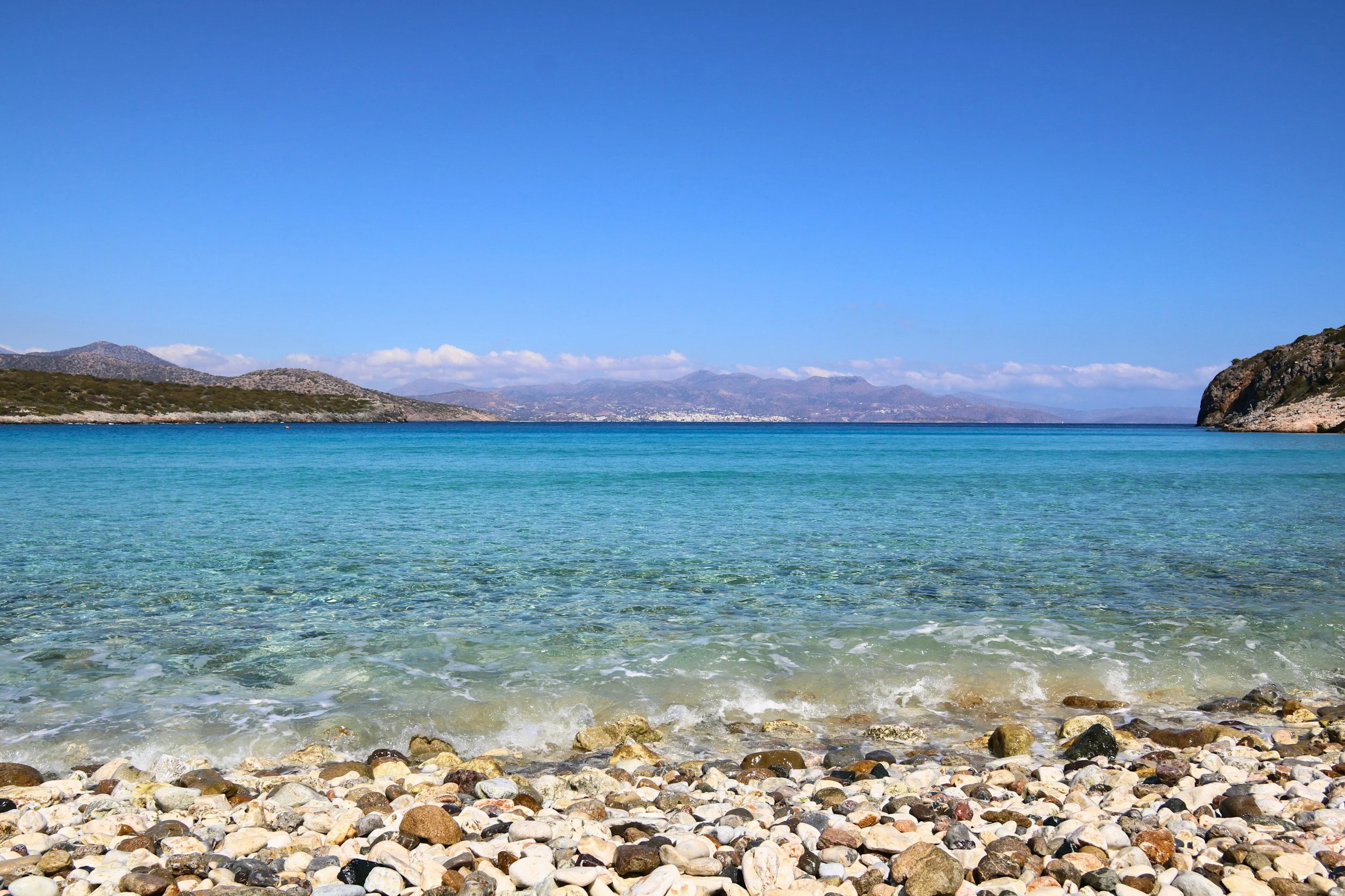 A rocky shoreline with small stones leading into a body of clear turquoise water. In the distance, there are hills and mountains with a mostly clear blue sky overhead.