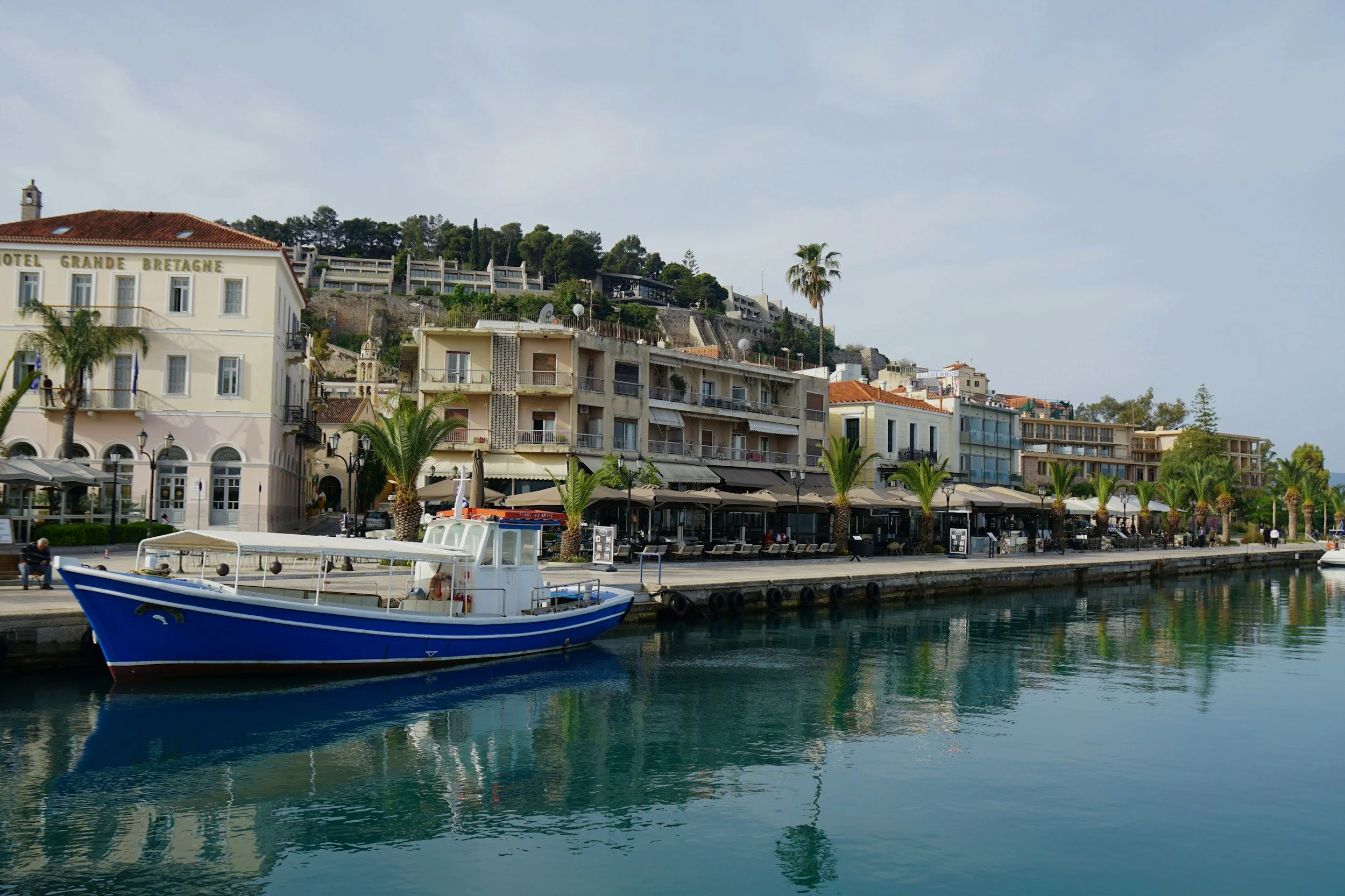 A marina with a blue and white boat docked along the water, palm trees, and buildings with balconies and red-tiled roofs on a cloudy day.