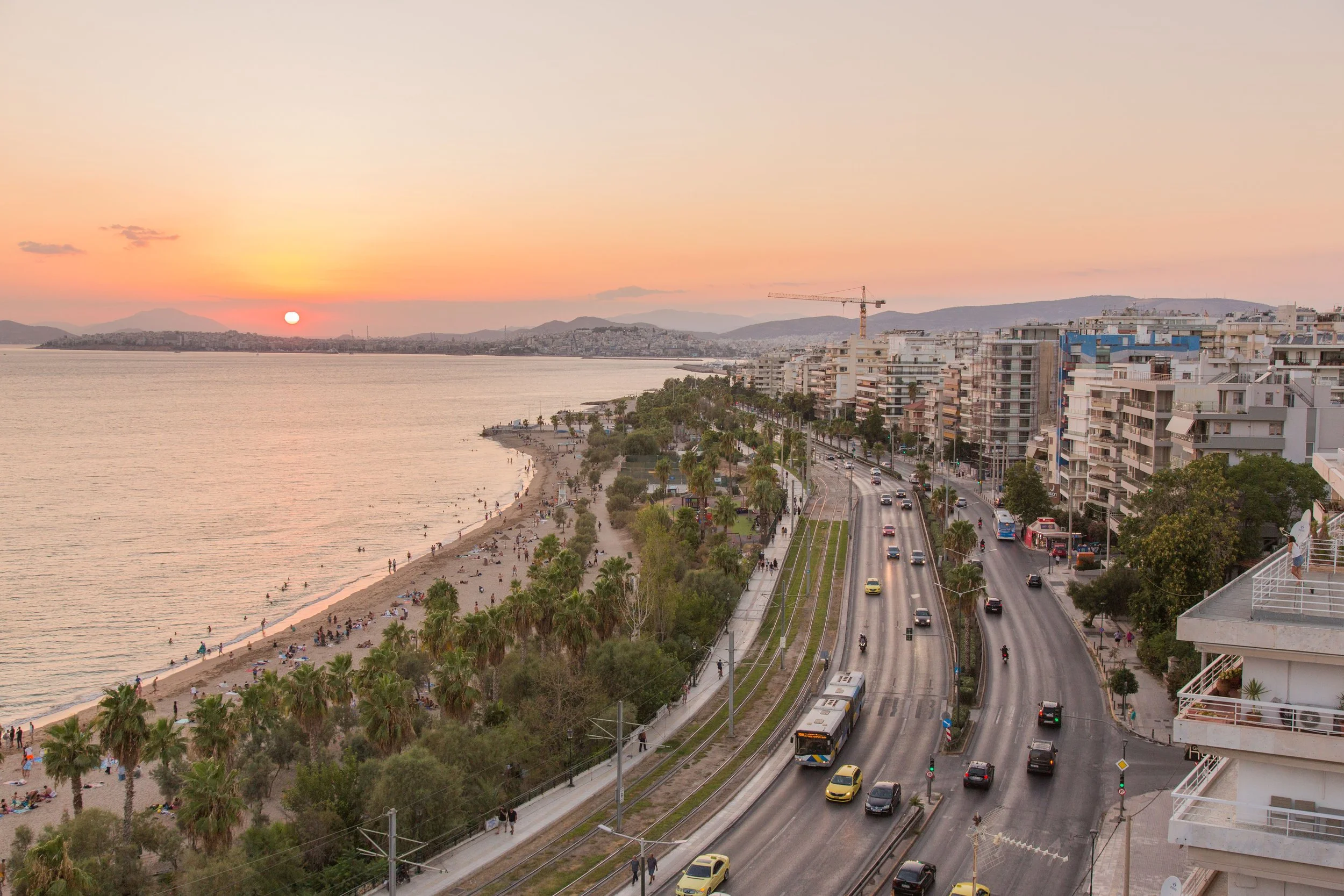 Sunset over a coastal city with a beach, palm trees, busy road, and modern buildings.