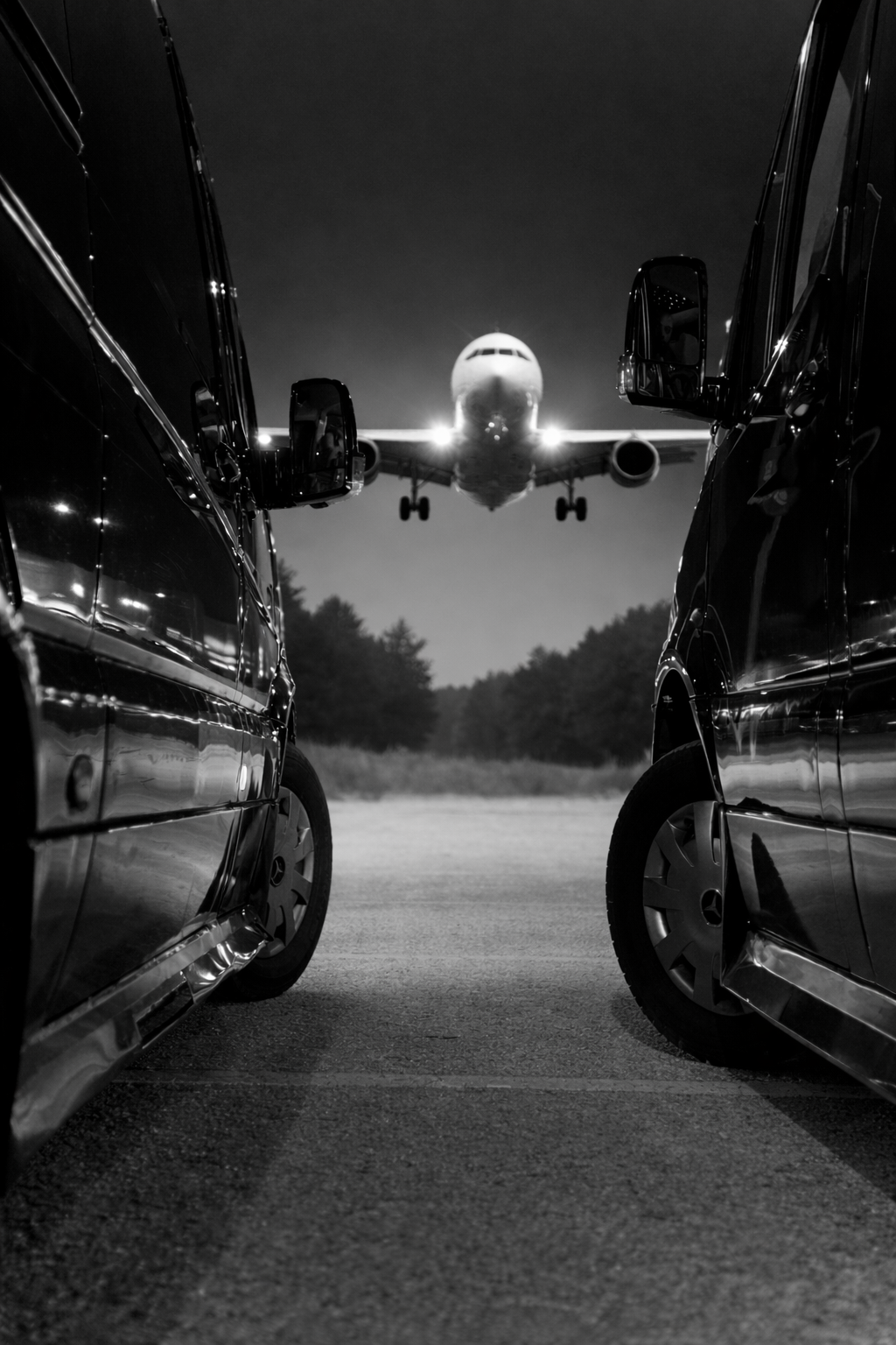Nighttime scene of an airplane approaching for landing, seen between two parked cars with highly reflective surfaces.