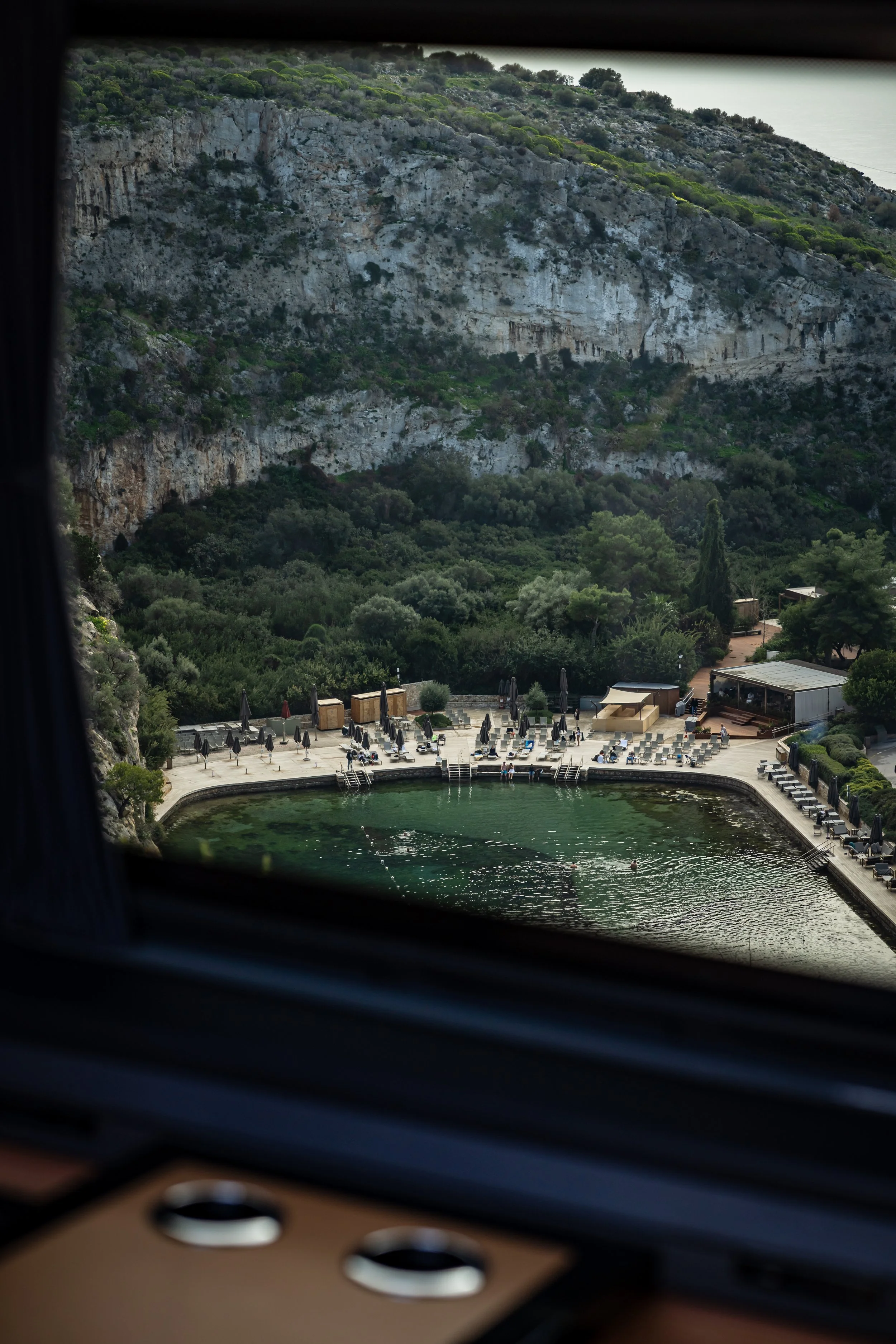 Photo of a rocky hillside with greenery and a small garden or park area below, viewed through a window with some blurred objects in the foreground.
