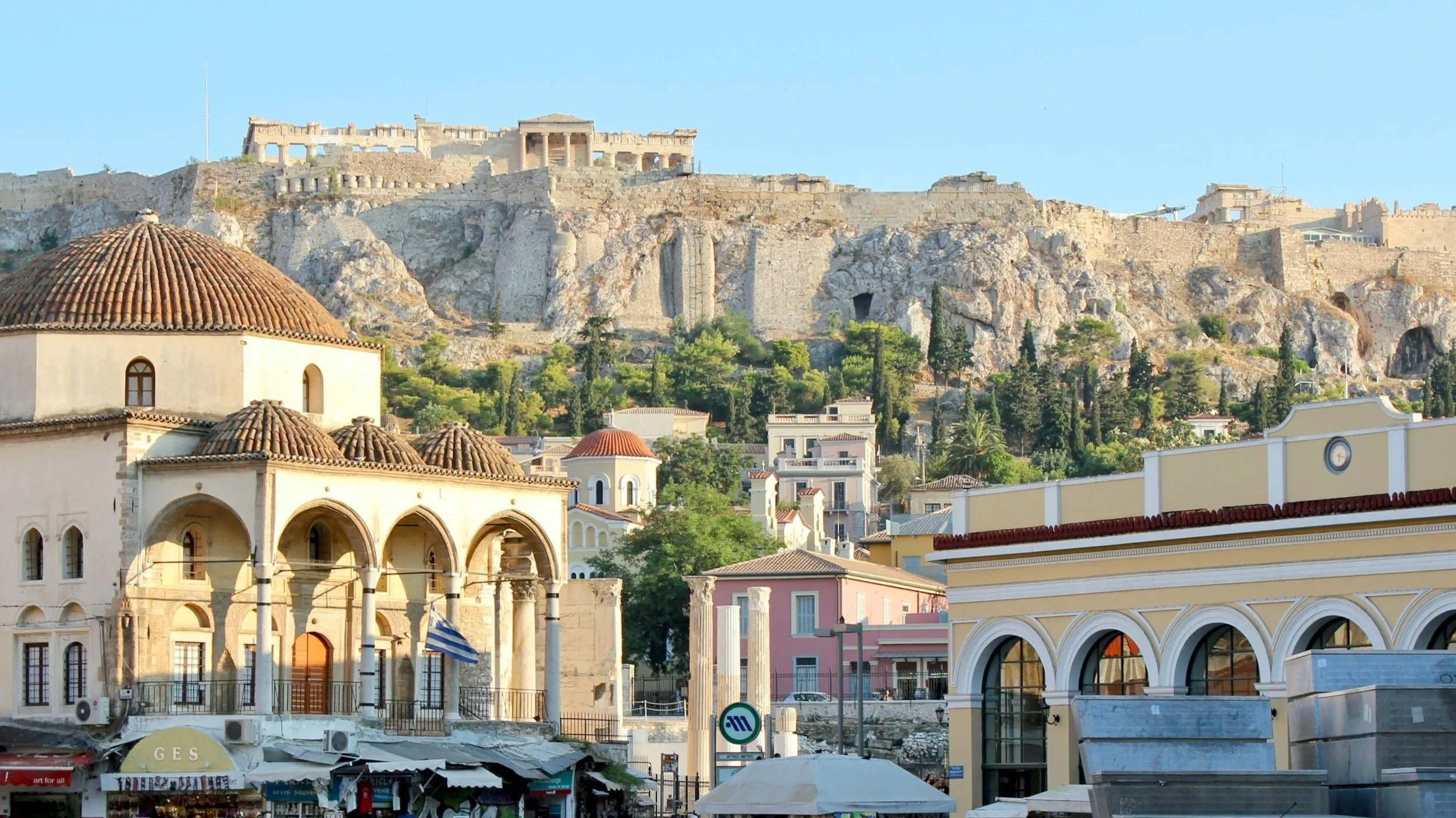 A view of Athens, Greece, showing historical buildings in the foreground, lush greenery in the middle, and the Acropolis on the rocky hilltop in the background, under a clear blue sky.