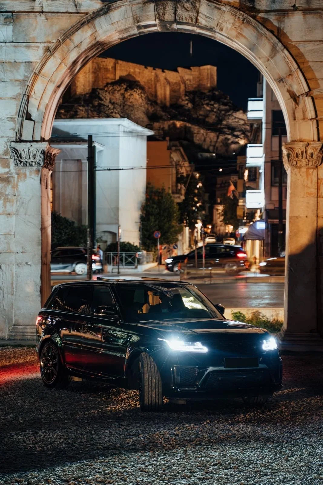 A black car with headlights on parked under an ancient stone archway at night, with a city street and rocky hilltop fortress visible through the arch.