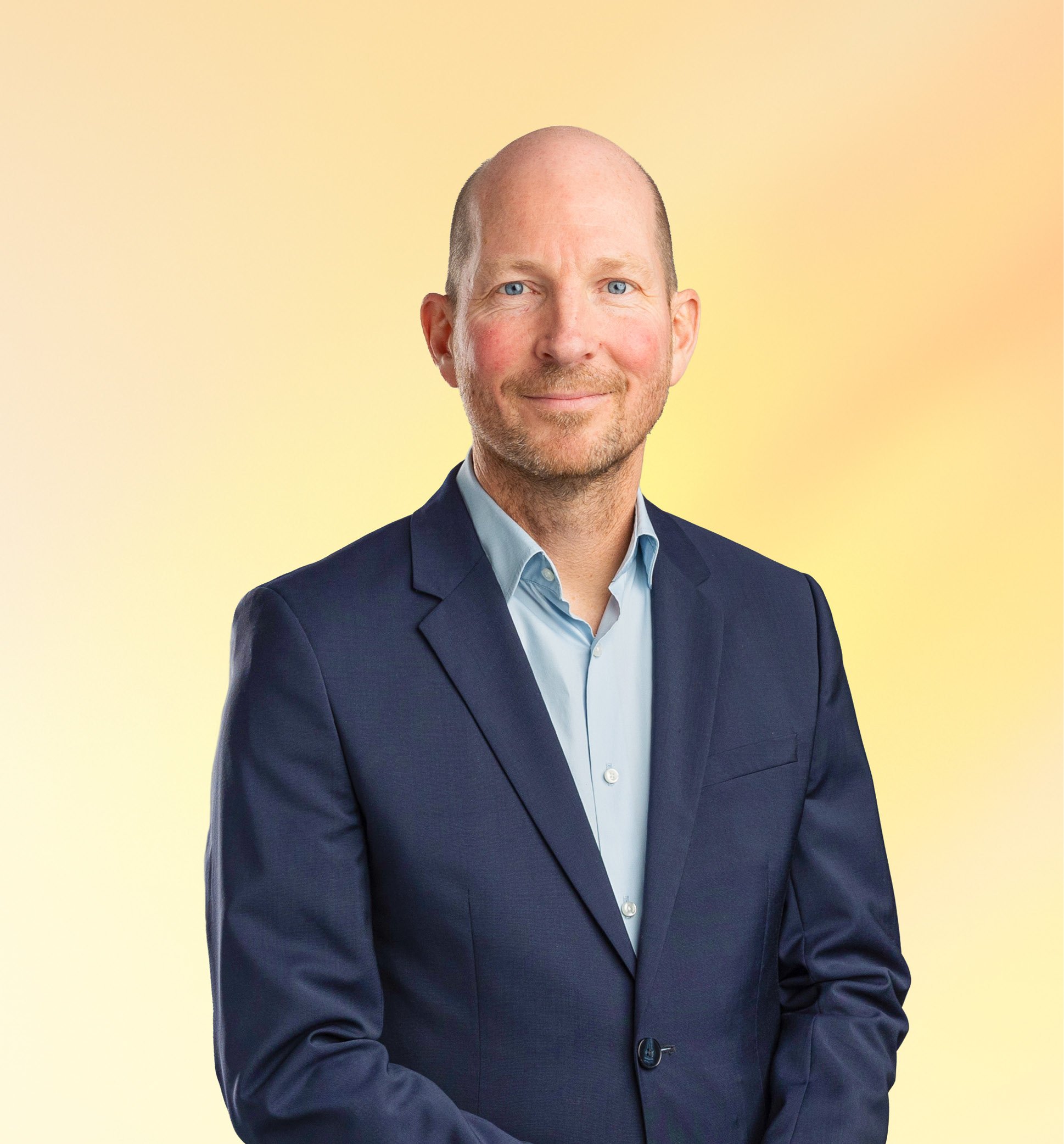 Man in een blauwe blazer met gekruiste armen, glimlachend, tegen een pastelkleurige achtergrond.