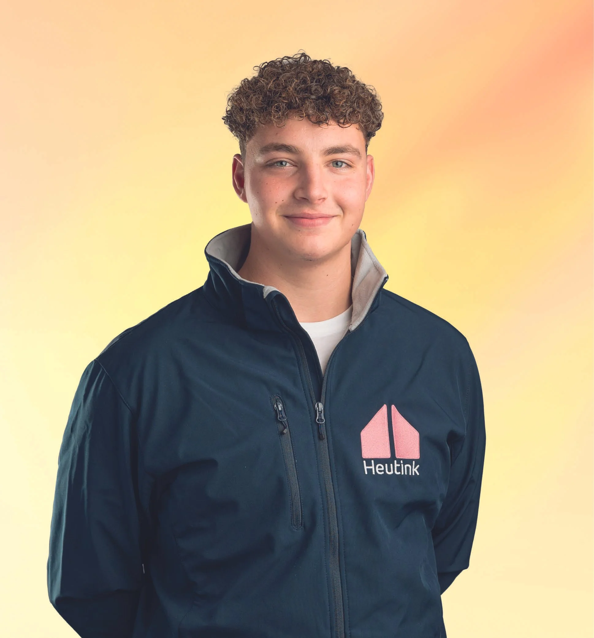 Portret van een jonge man met krullend haar, gekleed in een zwart jack met een logo en tekst 'Heutink', staand tegen een gele en roze achtergrond.