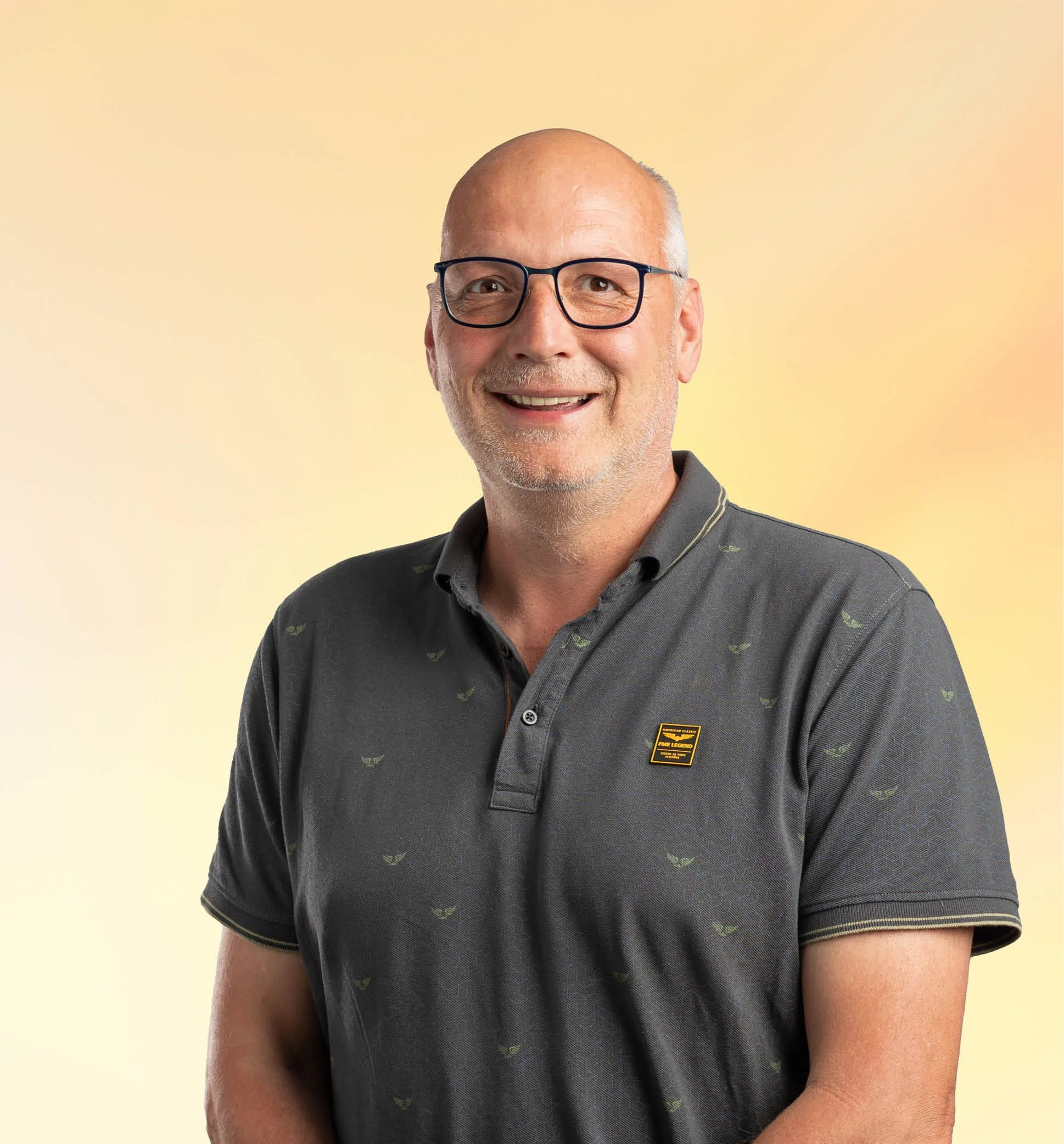 Een glimlachende man met een bril en een grijze polo met een klein geel en zwart logo, staand voor een lichthuidige achtergrond met een zachte kleurverloop.
