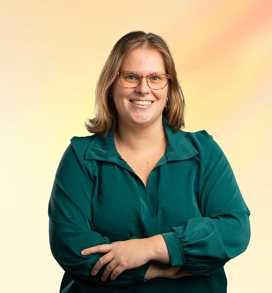 Vrouw met bruin haar, brillen, groene blouse, lachend met gekruiste armen voor een kleurige achtergrond.