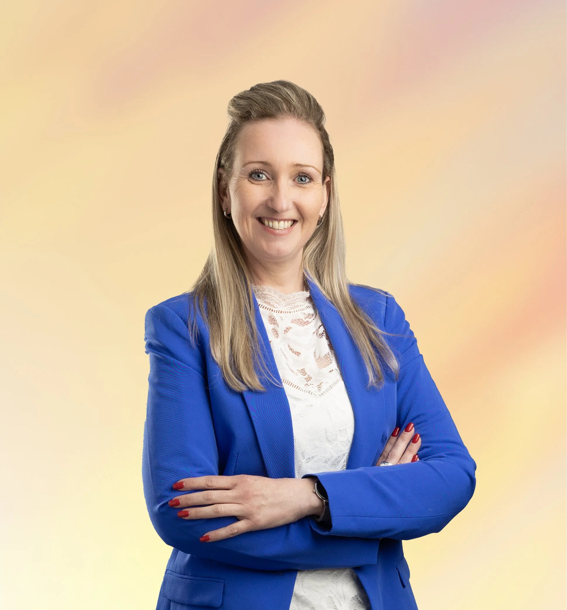 Een vrouw met blond haar, gekleed in een blauwe jas en een witte blouse, die glimlacht en haar armen gekruist heeft.