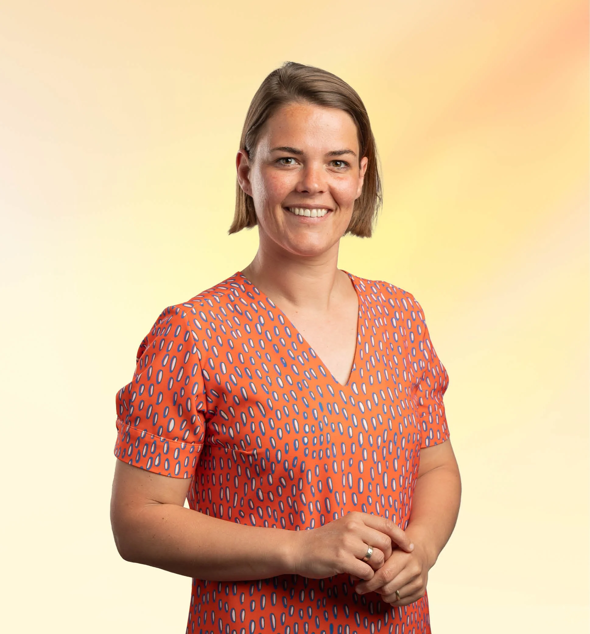Portret van een lachende vrouw met schouderlange bruin haar, gekleed in een oranje jurk met blauwe en witte patronen, standend tegen een lichte gele achtergrond.