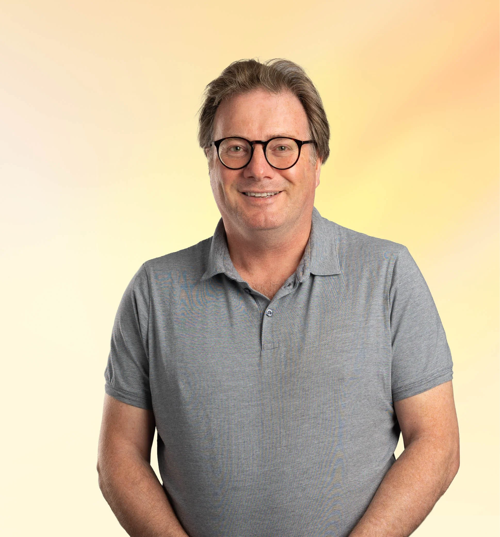 Man met bruin haar, bril en grijs polo, glimlachend voor een gele achtergrond.