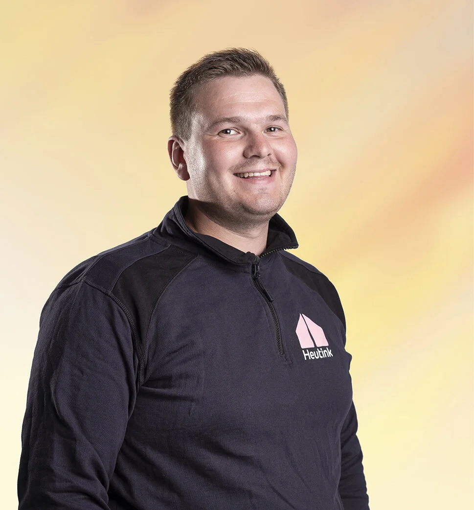 Een glimlachende man met kort, lichtbruin haar draagt een zwarte jas met logo en tekst 'Huitink' voor een pastel gekleurde achtergrond.