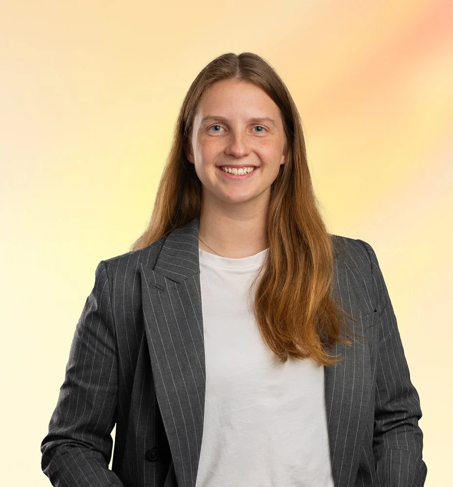 Vrouw met lang bruin haar, draag een zwart gestreept pak en witte t-shirt, glimlachend naar de camera, op een lichte, gekleurde achtergrond.