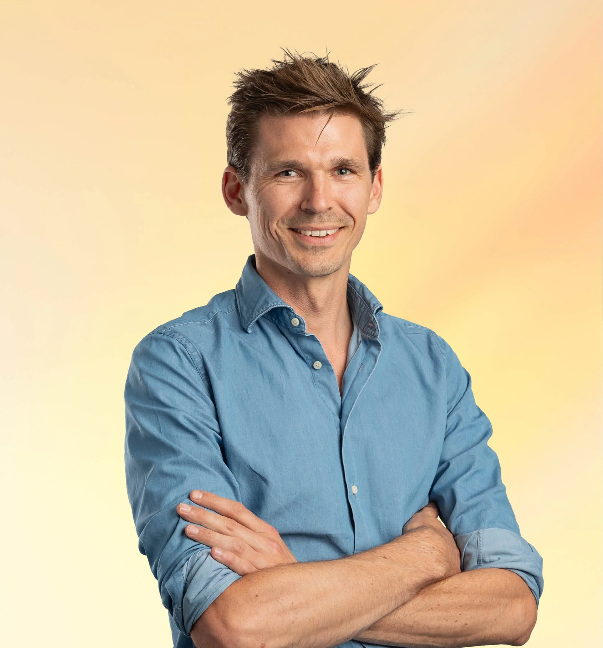 Portret van een lachende man met gevouwen armen, blauwe blouse, lichte achtergrond.