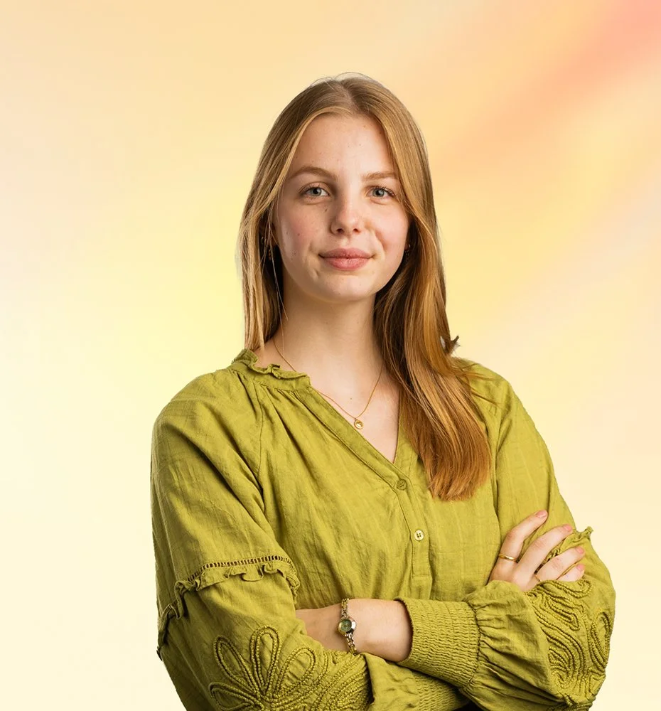 Een jonge vrouw met lang, roodbruin haar die een groene blouse draagt, staat met gekruiste armen voor een kleurrijke, lichte achtergrond.