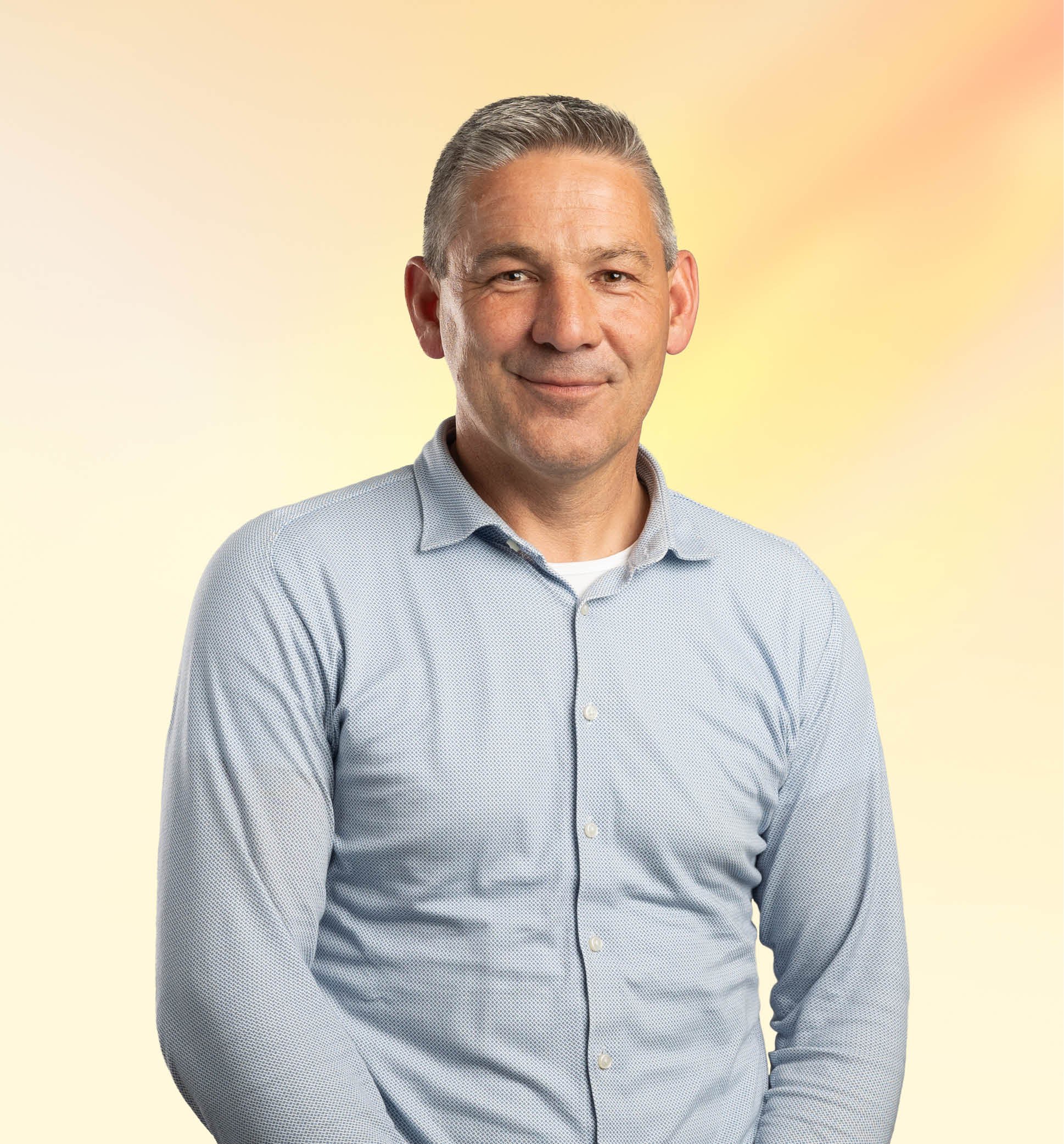 Portret van een man met kort, grijs haar en een glimlach, gekleed in een lichtblauw overhemd, staand voor een gele en roze gradient achtergrond.