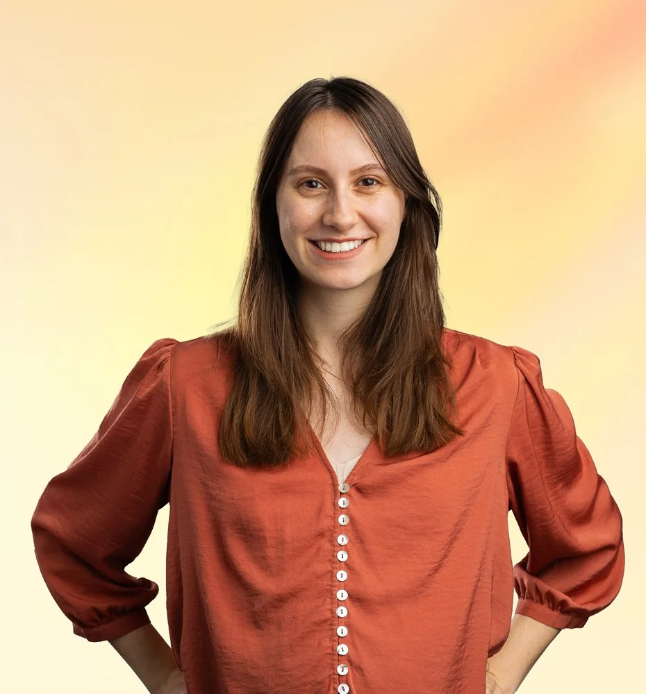 Lachende vrouw met bruin haar, draag een oranje blouse, poseert met handen op haar heupen voor een kleurrijke achtergrond.