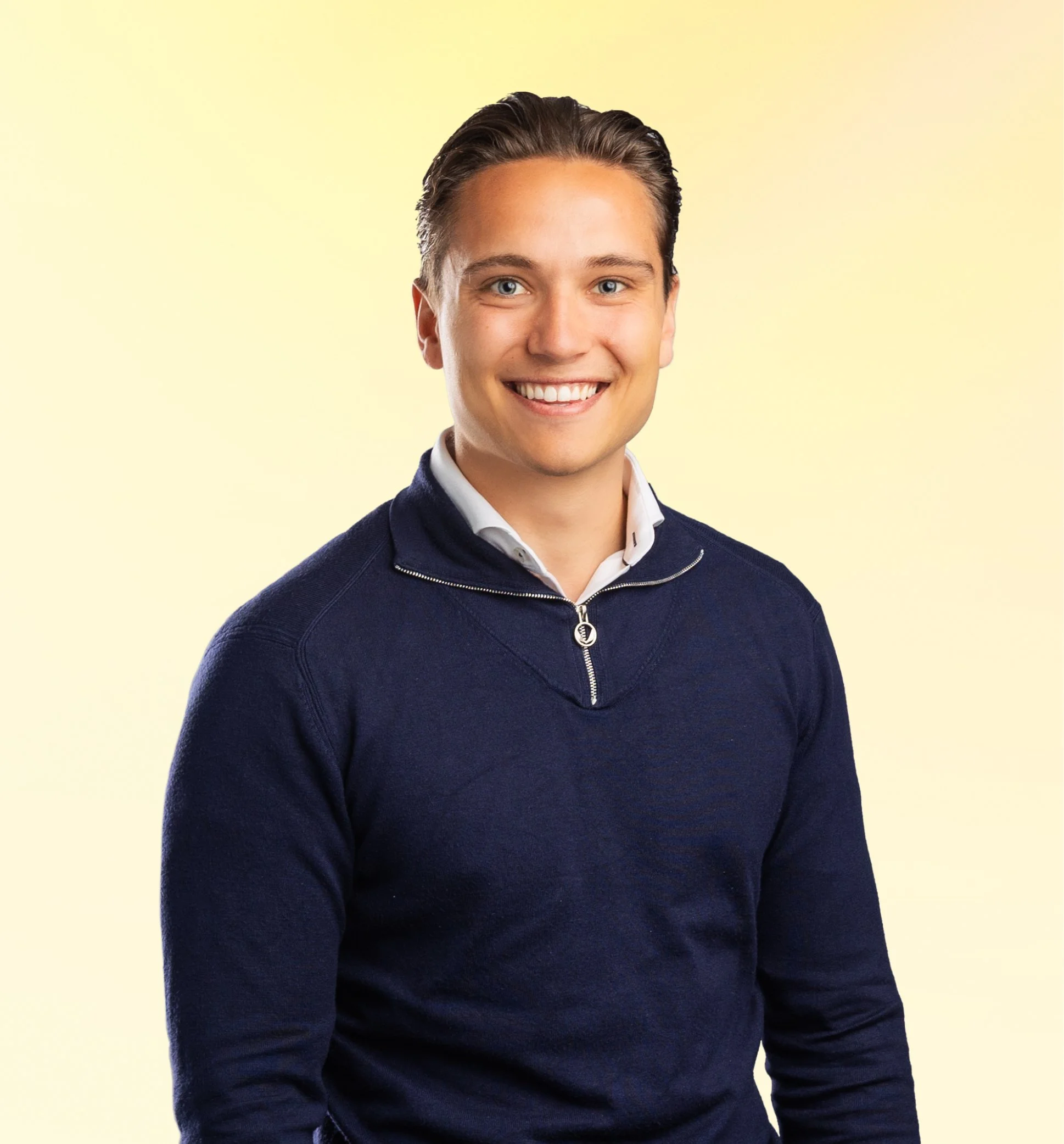 Portret van een lachende jonge man met donker haar, gekleed in een navyblauwe sweater met een witte blouse op een lichte achtergrond.